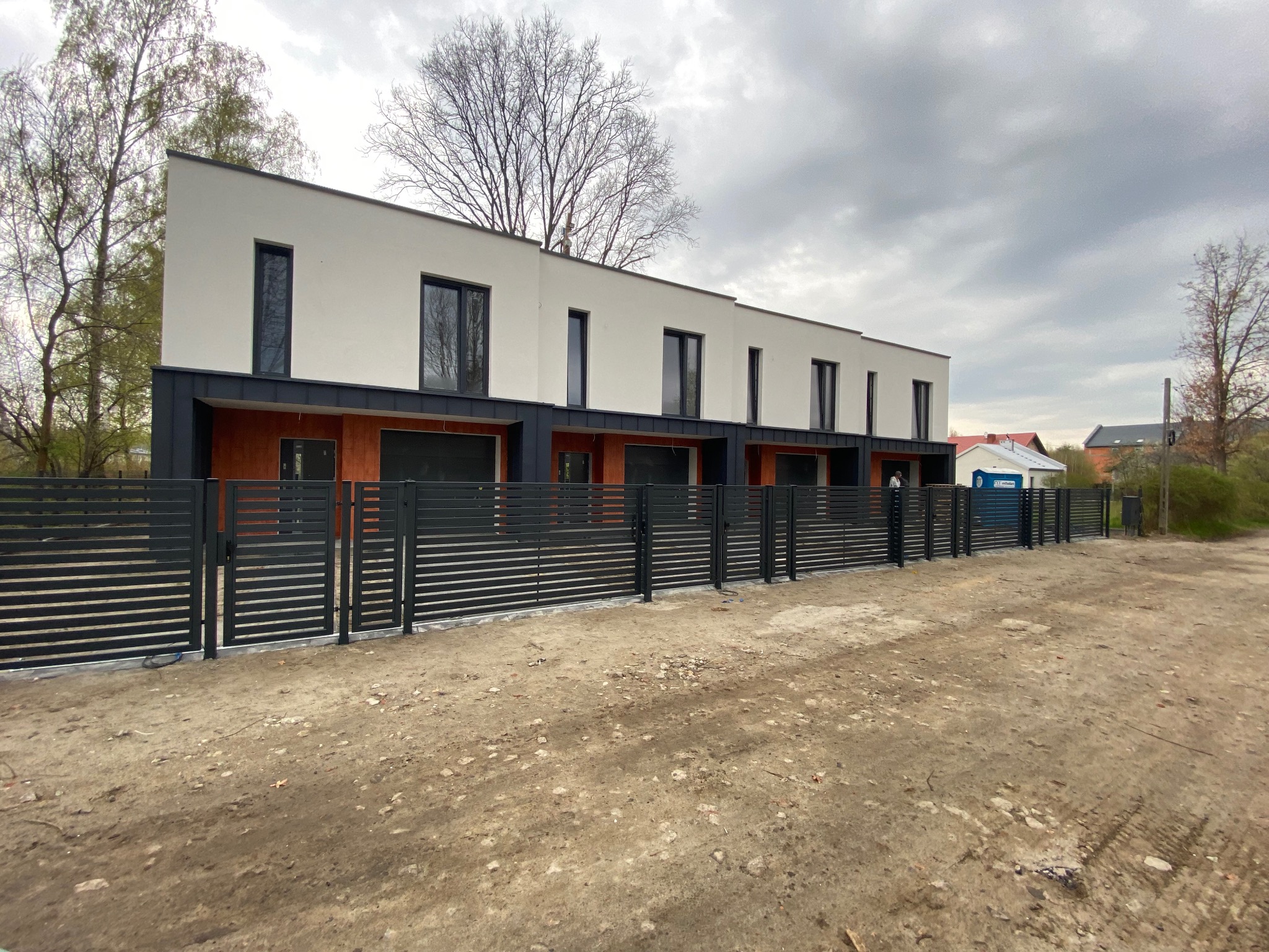 Szeregowe budynki jednorodzinne w stanie deweloperskim z elewacją w kolorze białym i czarnymi oknami, ogrodzone nowoczesnym, poziomym ogrodzeniem z metalowych sztachet na tle pochmurnego nieba.