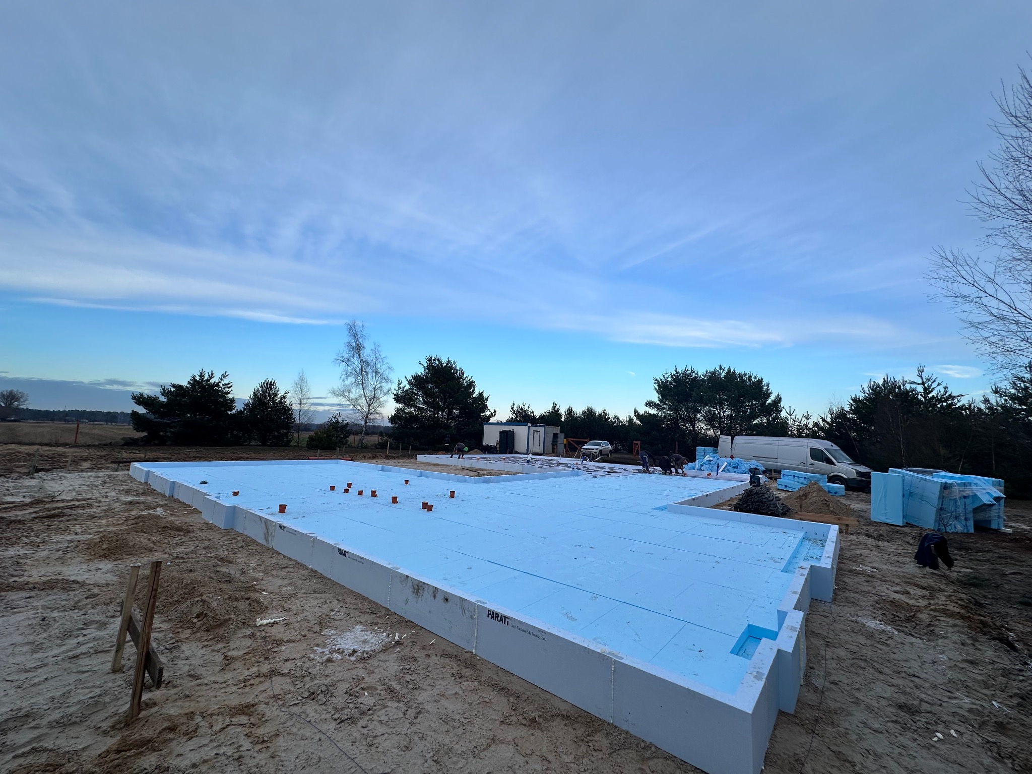 Ocieplona płyta fundamentowa z widocznymi elementami izolacji termicznej, na tle piaszczystego terenu budowy i częściowo zachmurzonego nieba.