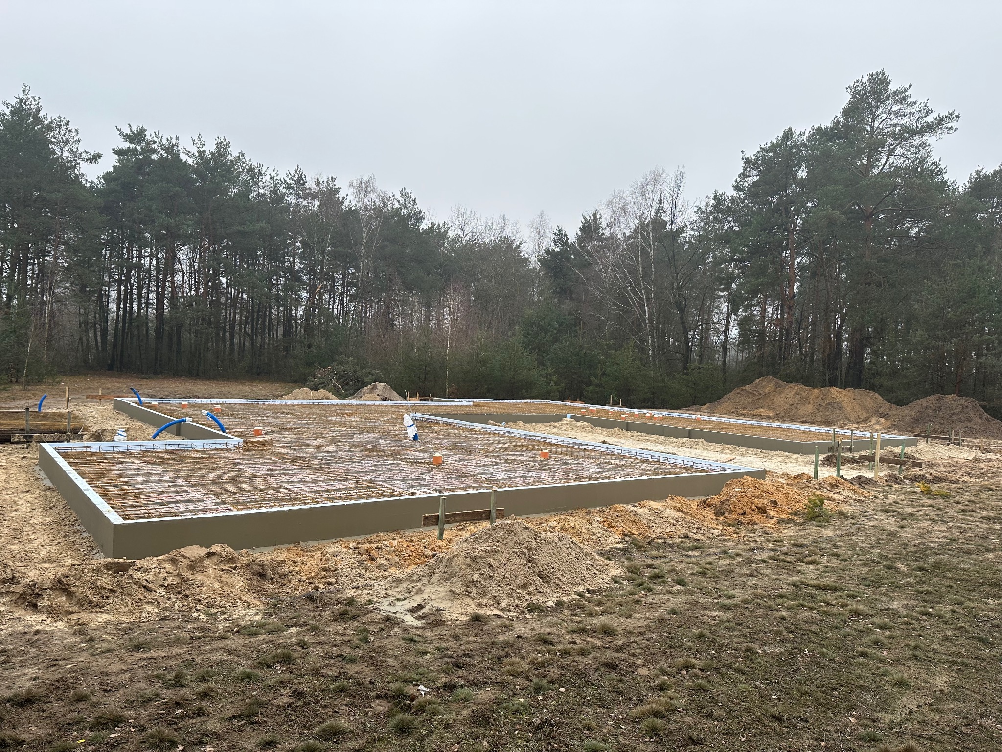 Zbrojona płyta fundamentowa z izolacją obwodową na działce z widocznym lasem w tle, przygotowana do dalszych prac budowlanych.