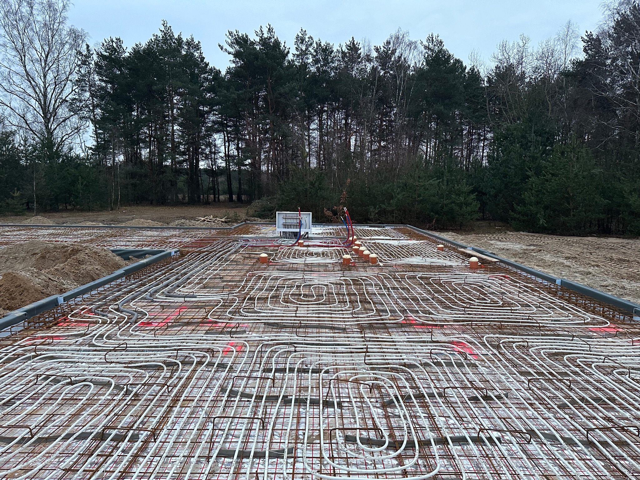Zbrojenie ocieplanej płyty fundamentowej z systemem ogrzewania podłogowego, widoczna instalacja elektryczna i teren budowy z drzewami w tle.
