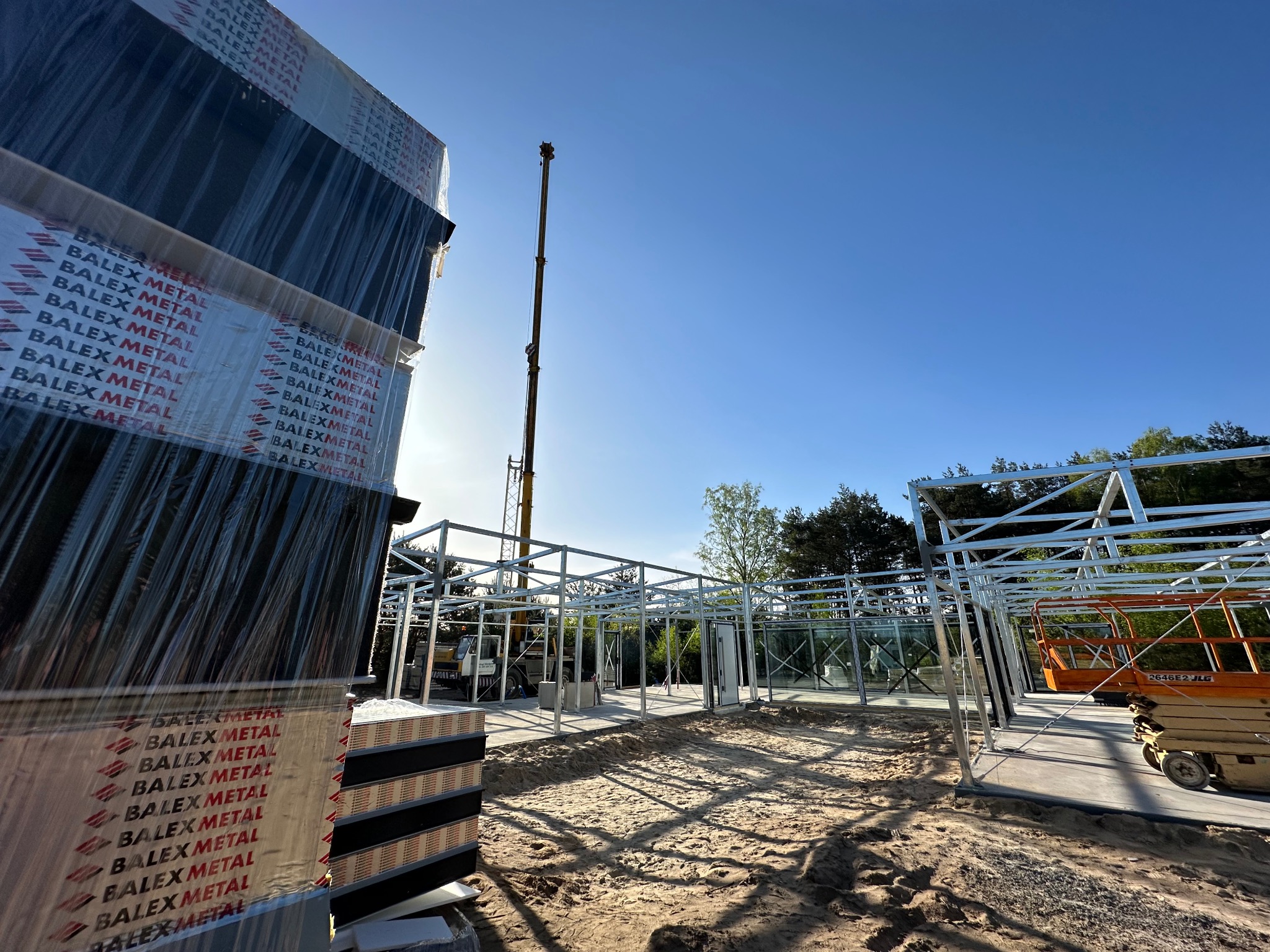 Konstrukcja domu na szkielecie stalowym w trakcie montażu, widoczne przygotowane pakiety płyt Balex Metal, żuraw budowlany w tle i podnośnik koszowy.