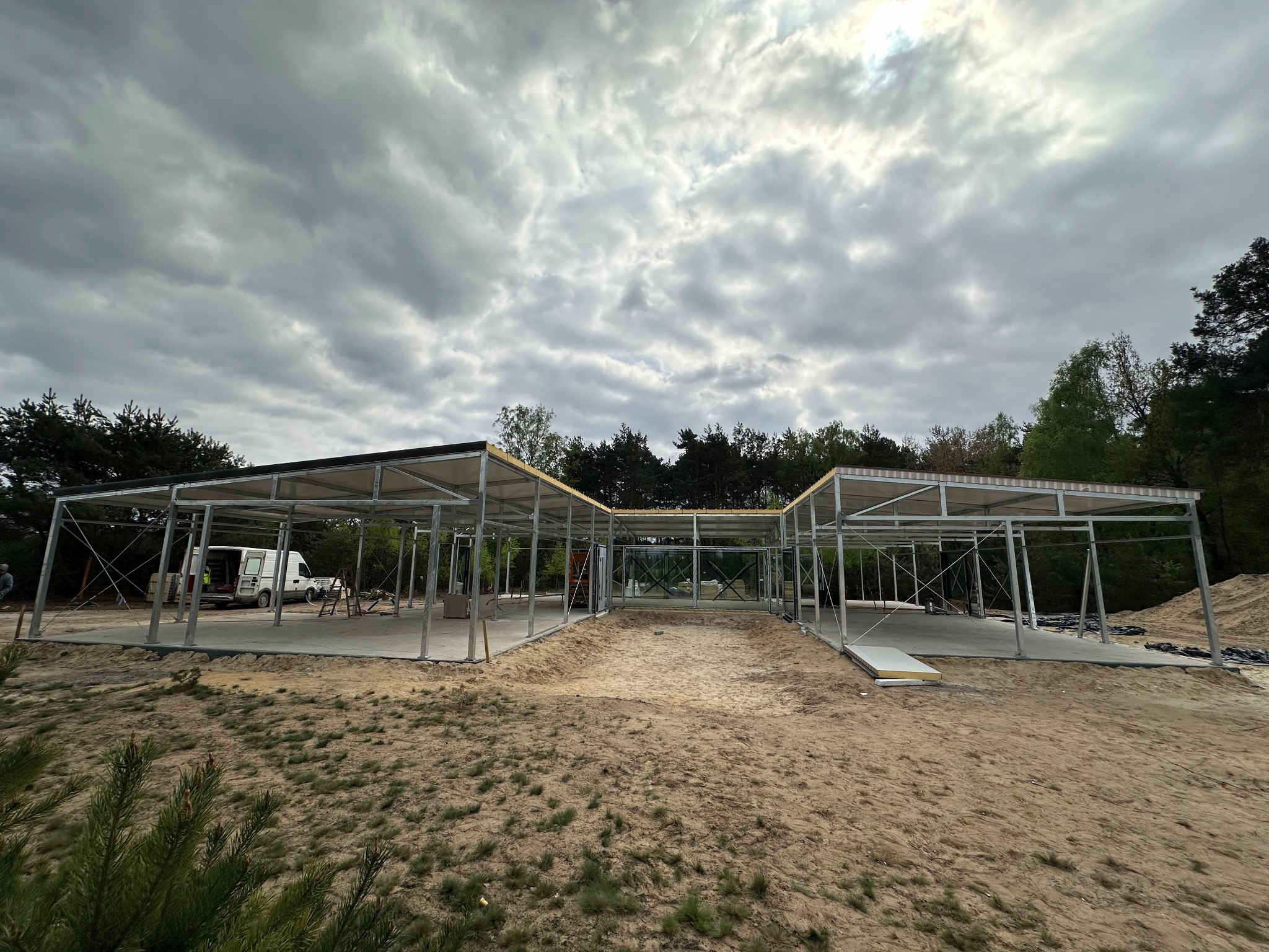 Konstrukcja szkieletowa budynku z zamontowanym dachem, widoczny van z otwartymi drzwiami w tle, teren piaszczysty, pochmurne niebo.