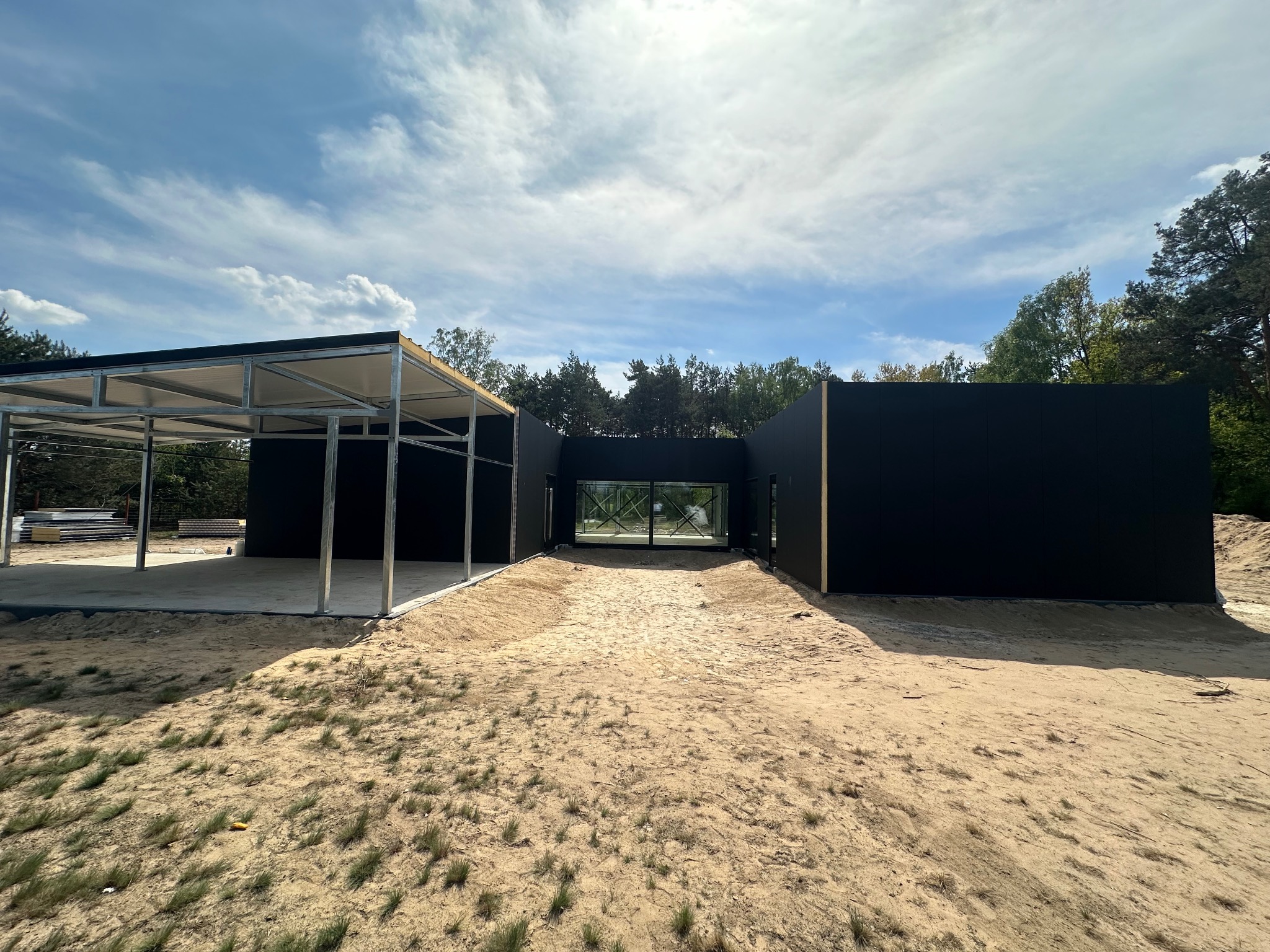 Nowoczesny dom modułowy w trakcie budowy z widoczną stalową konstrukcją carportu i czarną elewacją, teren piaszczysty.
