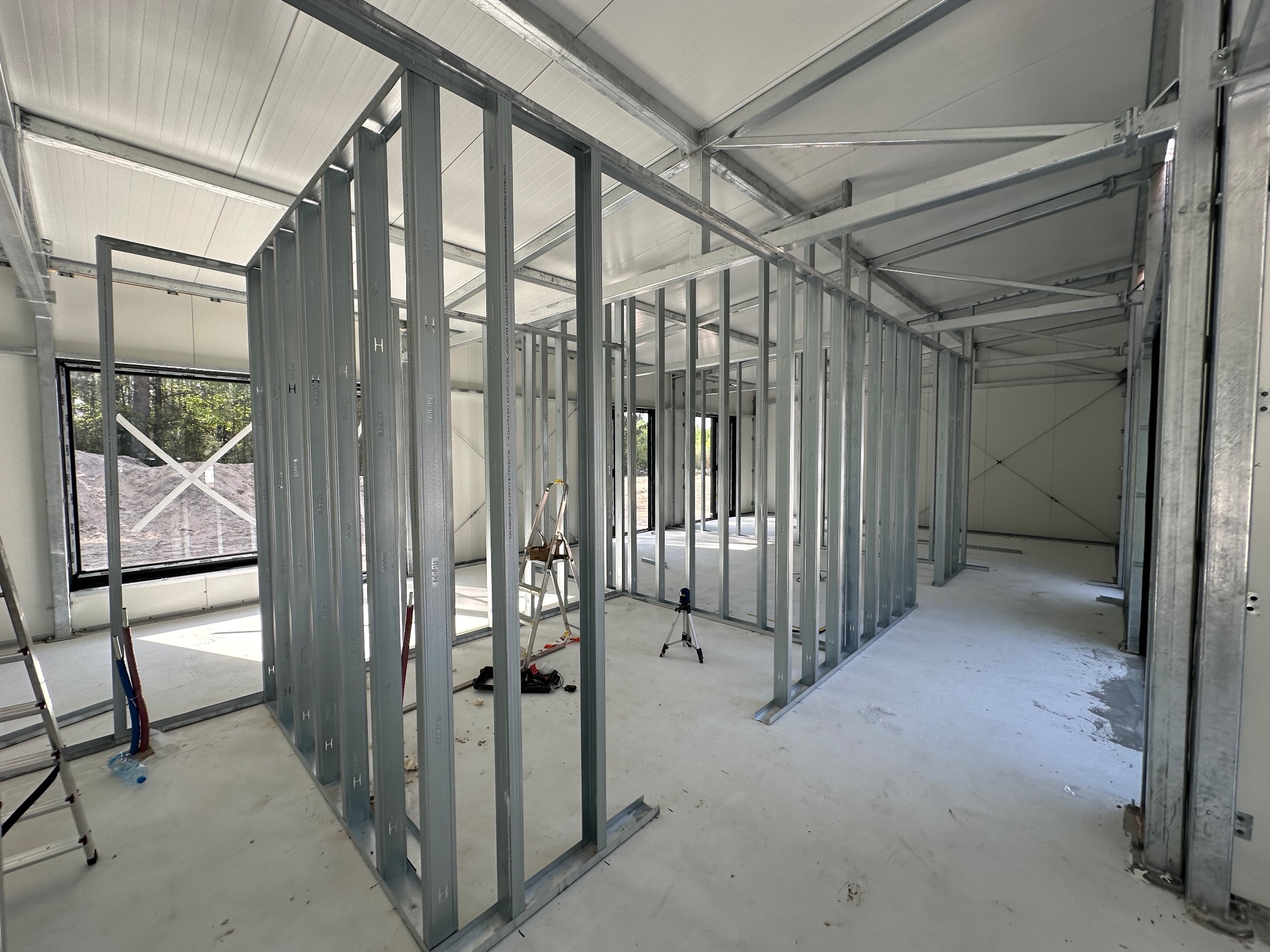 Wnętrze budynku w trakcie budowy, widoczne stalowe profile konstrukcyjne ścian działowych, poziomica laserowa na statywie, drabina i narzędzia na podłodze.