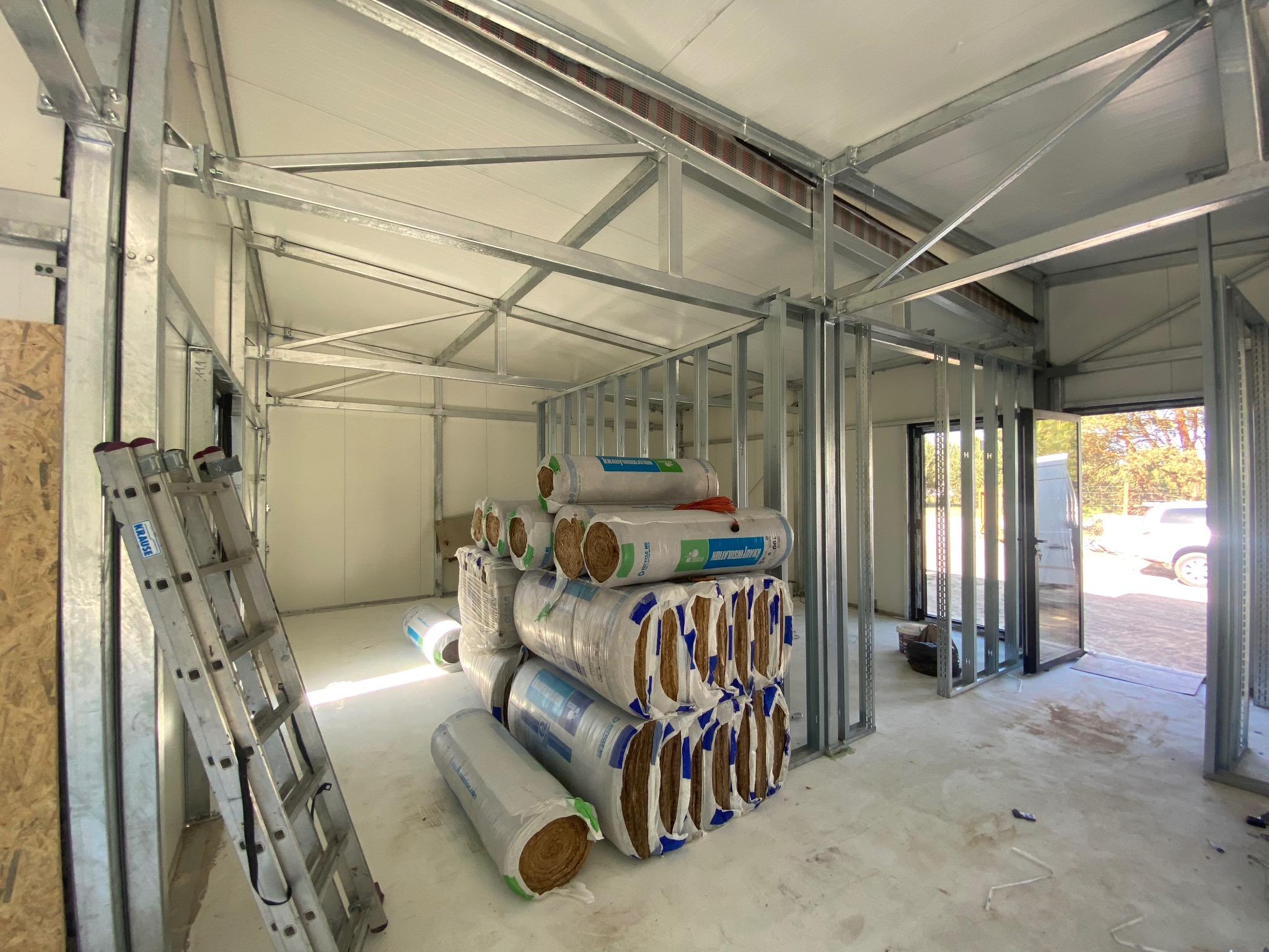 Wnętrze budynku w trakcie budowy, widoczny szkielet stalowy ścian i dachu, białe panele ścienne, stos izolacji z wełny mineralnej w folii oraz aluminiowa drabina oparta o ścianę.