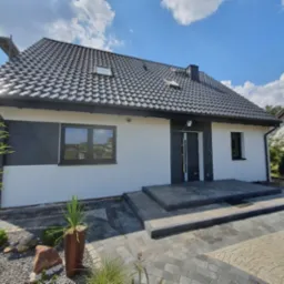 Nowoczesny dom jednorodzinny z ciemną dachówką i elewacją w kolorze biało-grafitowym, widok z poziomu brukowanego podjazdu z elementami dekoracyjnymi i roślinnością.