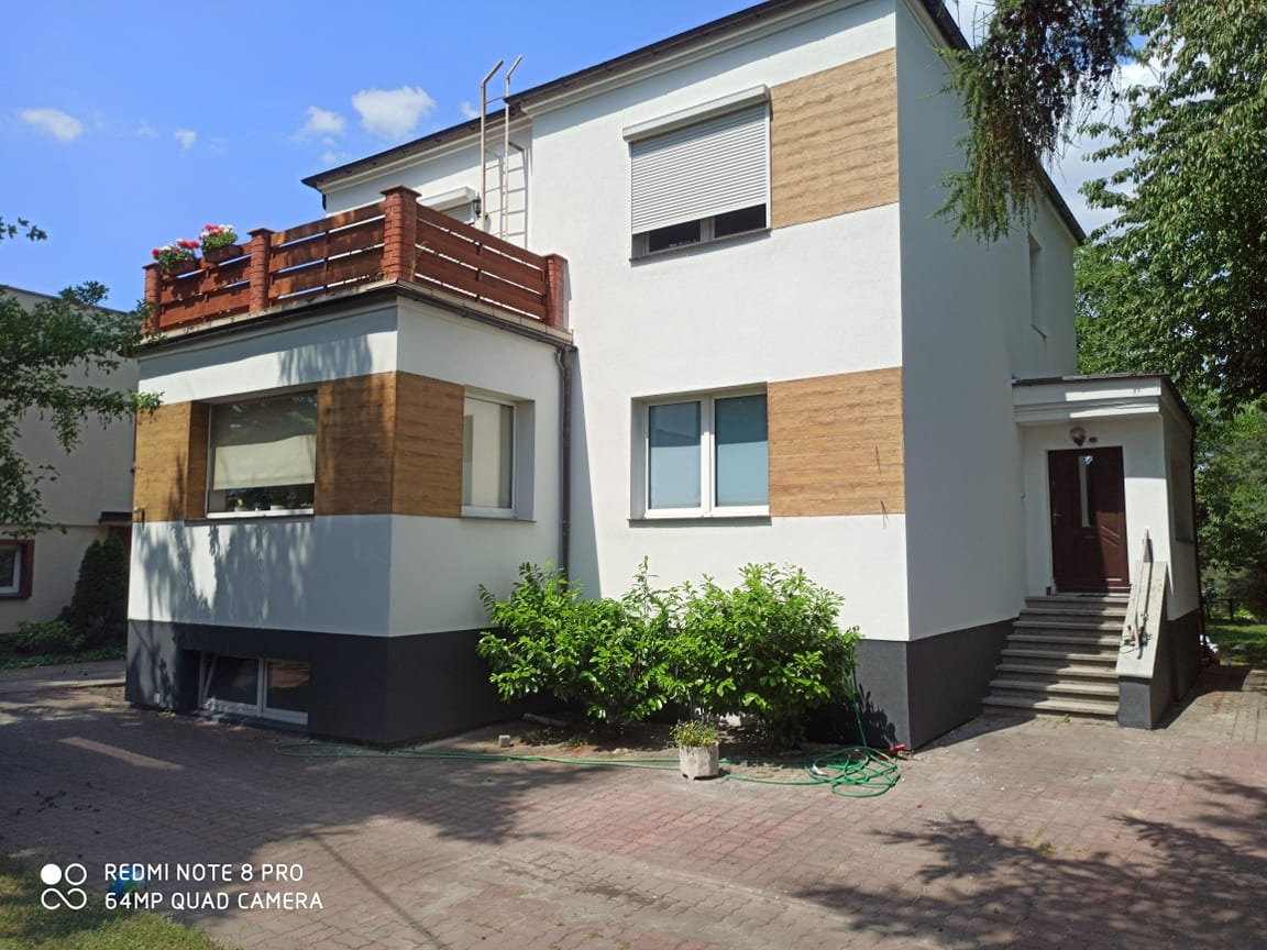 Dwupiętrowy dom z balkonem, białą elewacją i poziomymi pasami imitującymi drewno, widok z zewnątrz w słoneczny dzień.