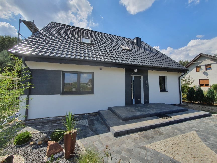 Nowoczesny dom jednorodzinny z ciemną dachówką i elewacją w kolorze biało-grafitowym, widok z poziomu brukowanego podjazdu z elementami dekoracyjnymi i roślinnością.