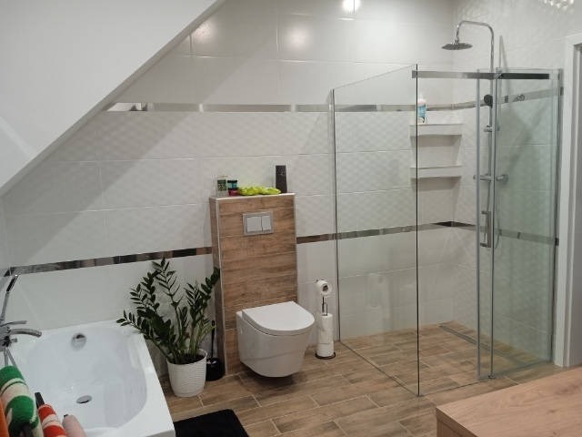 Nowoczesna łazienka na poddaszu z kabiną prysznicową ze szklanymi ścianami, białą wanną, toaletą podwieszaną na drewnianej płycie i rośliną doniczkową.