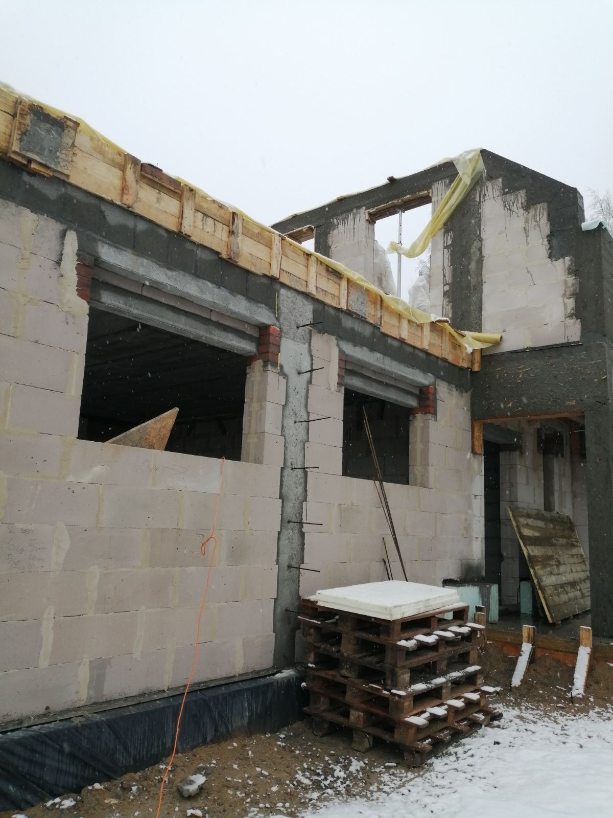 Budowa domu w trakcie zimy: surowe ściany z betonu komórkowego, drewniane szalunki nad otworami okiennymi, widoczne zbrojenia, ocieplenie fundamentów i paleta z materiałami budowlanymi w śniegu.