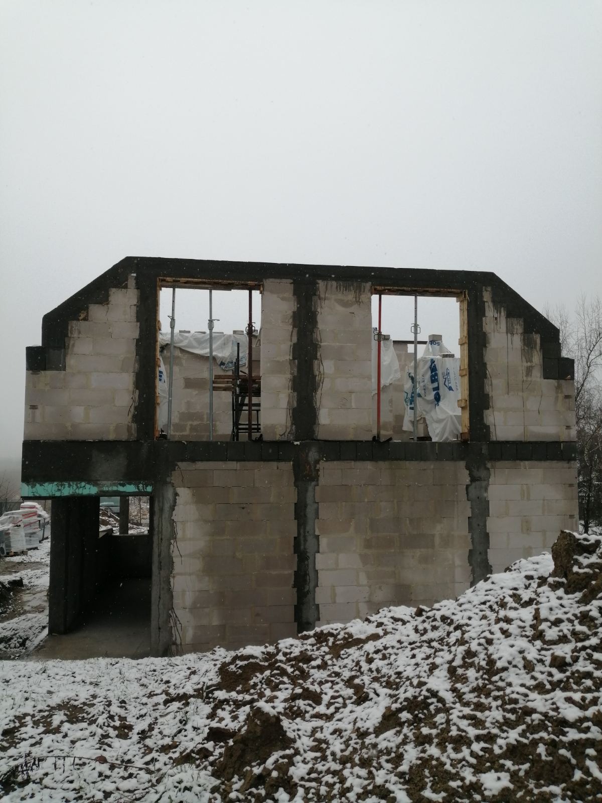 Surowy stan budynku w trakcie budowy, widoczne ściany z pustaków, otwory okienne zabezpieczone folią, zima, śnieg na ziemi.