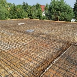 Zbrojenie stropu z siatki stalowej na deskowaniu, widok z góry, przygotowane do zalania betonem, w tle zieleń drzew i fragment budynku.