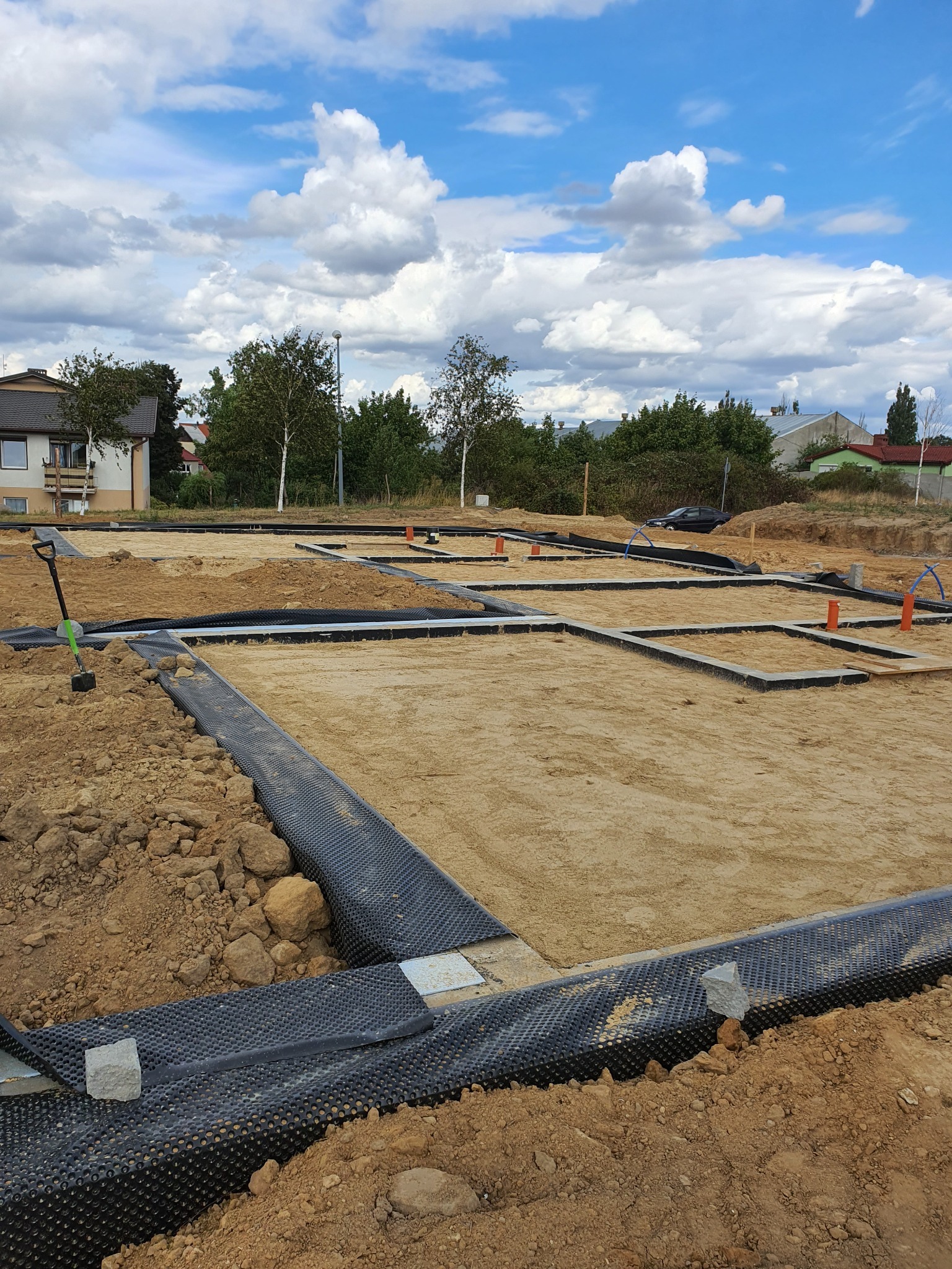 Przygotowanie fundamentów pod budowę domu: wykopane rowy zalane betonem, zabezpieczone folią izolacyjną, widoczna łopata i rury instalacyjne.