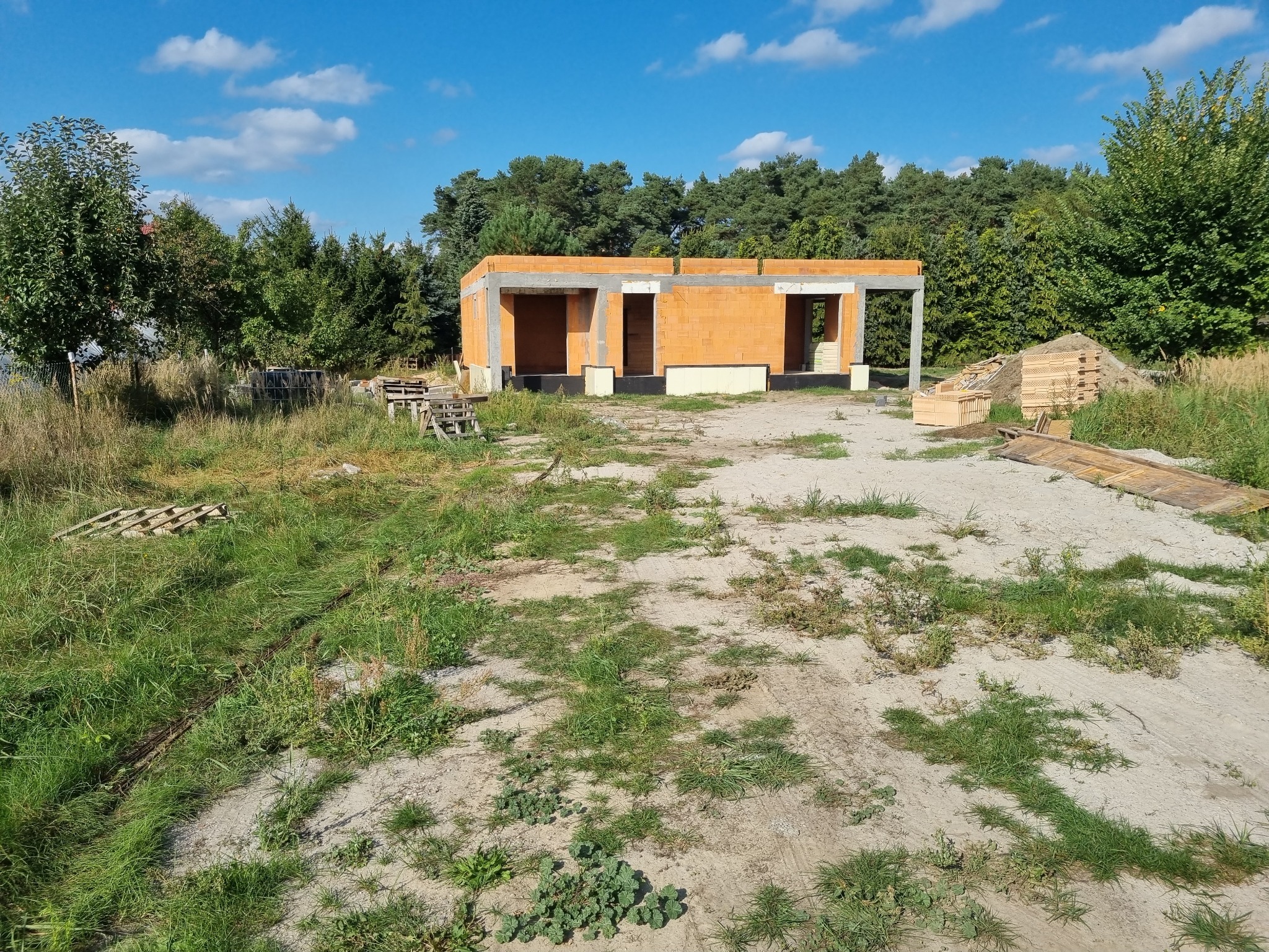 Surowy stan budynku jednorodzinnego z czerwonej cegły, widoczne fundamenty i ściany nośne, teren wokół niezagospodarowany, w tle drzewa i błękitne niebo.