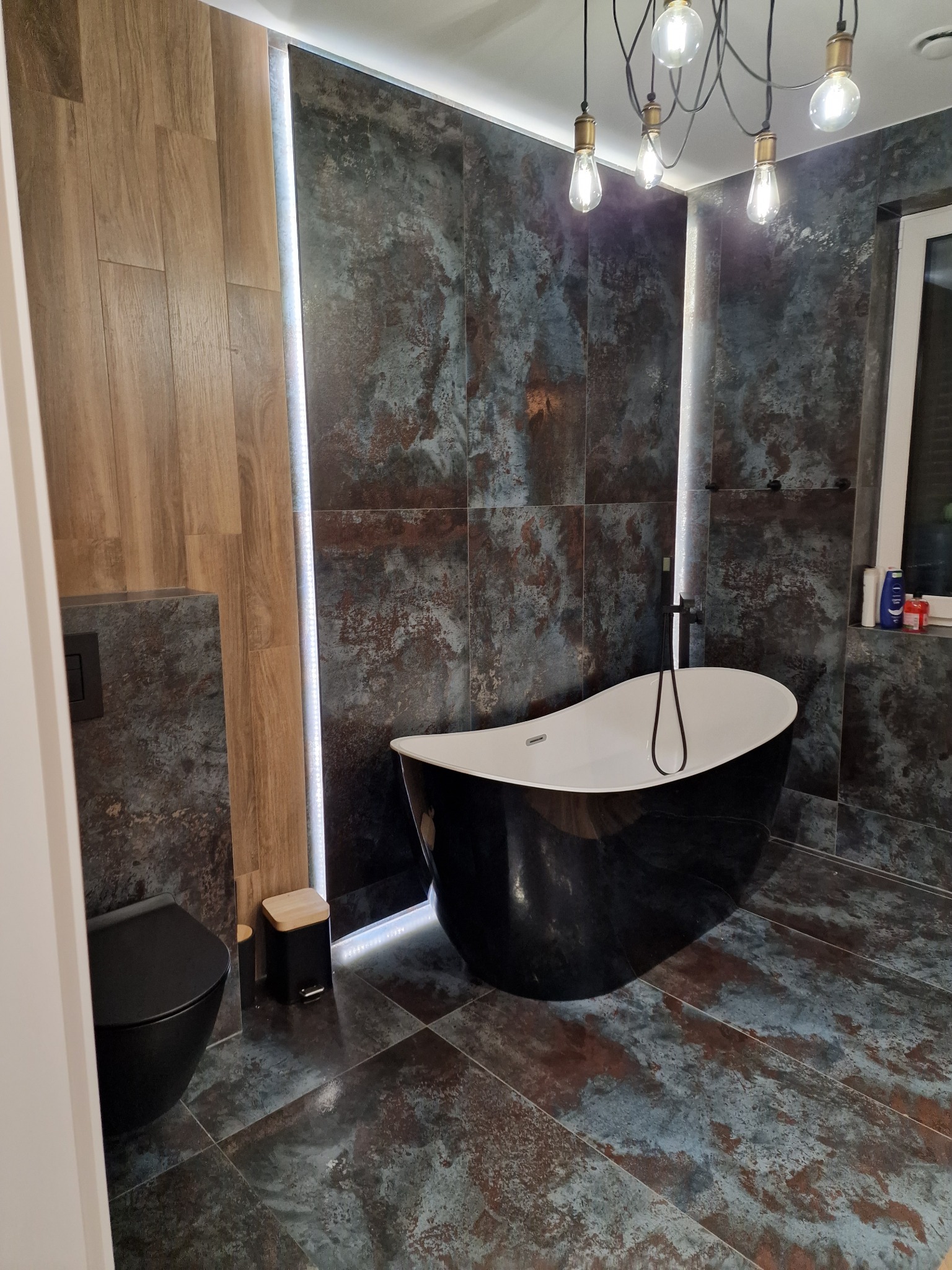 Nowoczesna łazienka z wolnostojącą czarną wanną, toaletą w kolorze czarnym i płytkami imitującymi rdzę, oświetlona dekoracyjnymi żarówkami Edison.