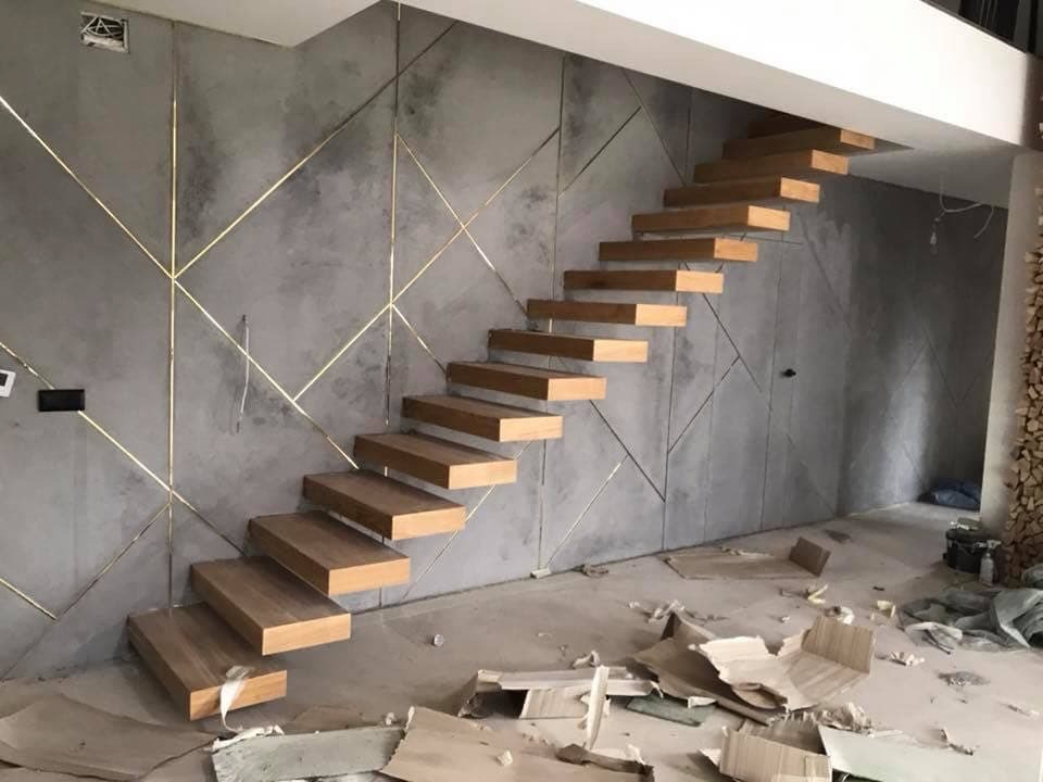 Nowoczesne, drewniane schody wiszące zamontowane przy szarej ścianie z geometrycznym wzorem z cienkich, złotych listew. Widoczne ślady prac wykończeniowych na podłodze.