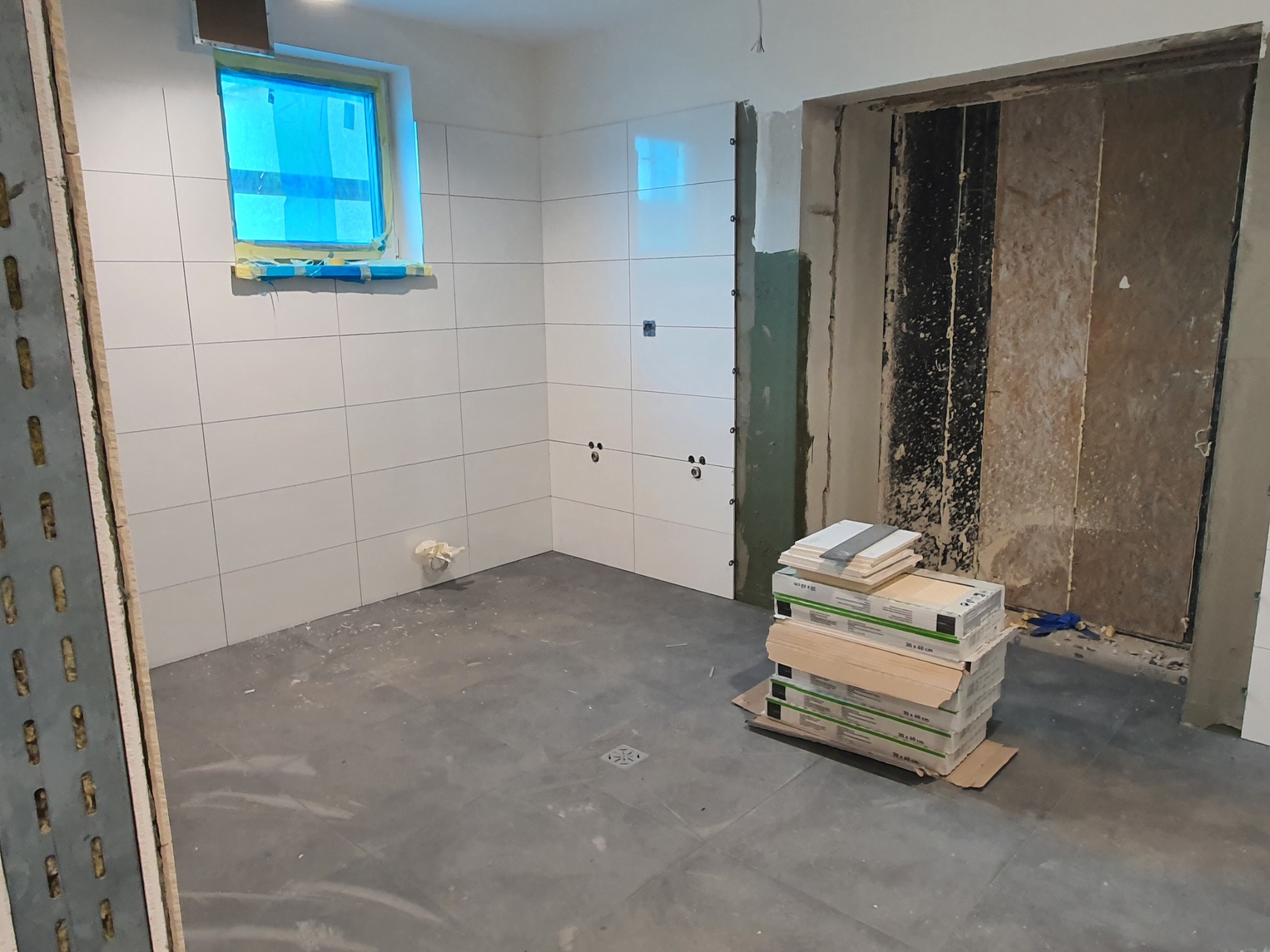 Wnętrze w trakcie remontu: ułożone białe płytki na ścianach, okno zabezpieczone folią, przygotowane paczki z płytkami podłogowymi, widoczny fragment konstrukcji ściany działowej.