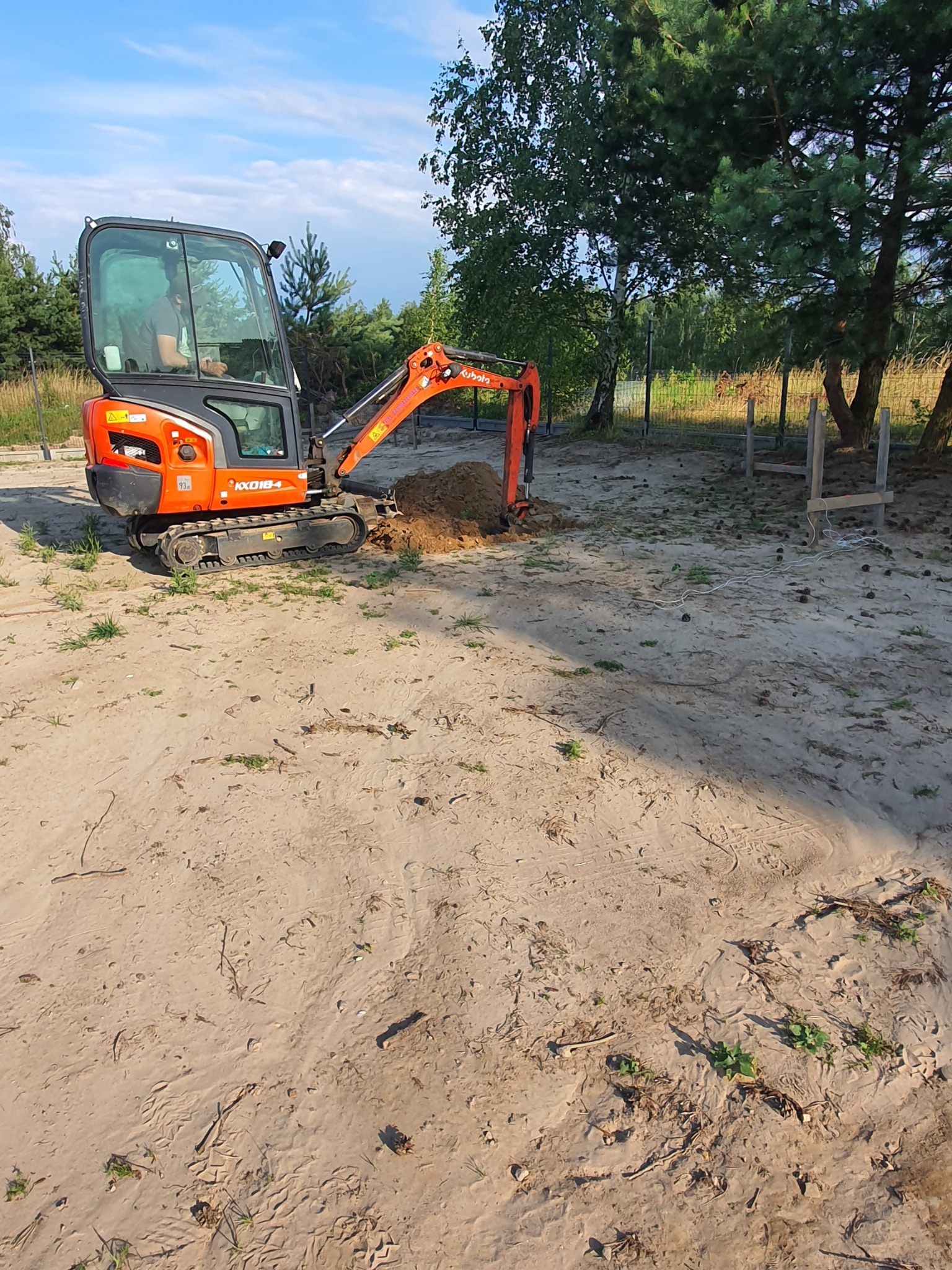 Pomarańczowa mini-koparka Kubota KX018-4 z operatorem, wykonująca wykop ziemny na piaszczystym terenie z widocznymi drzewami i elementami ogrodzenia w tle.