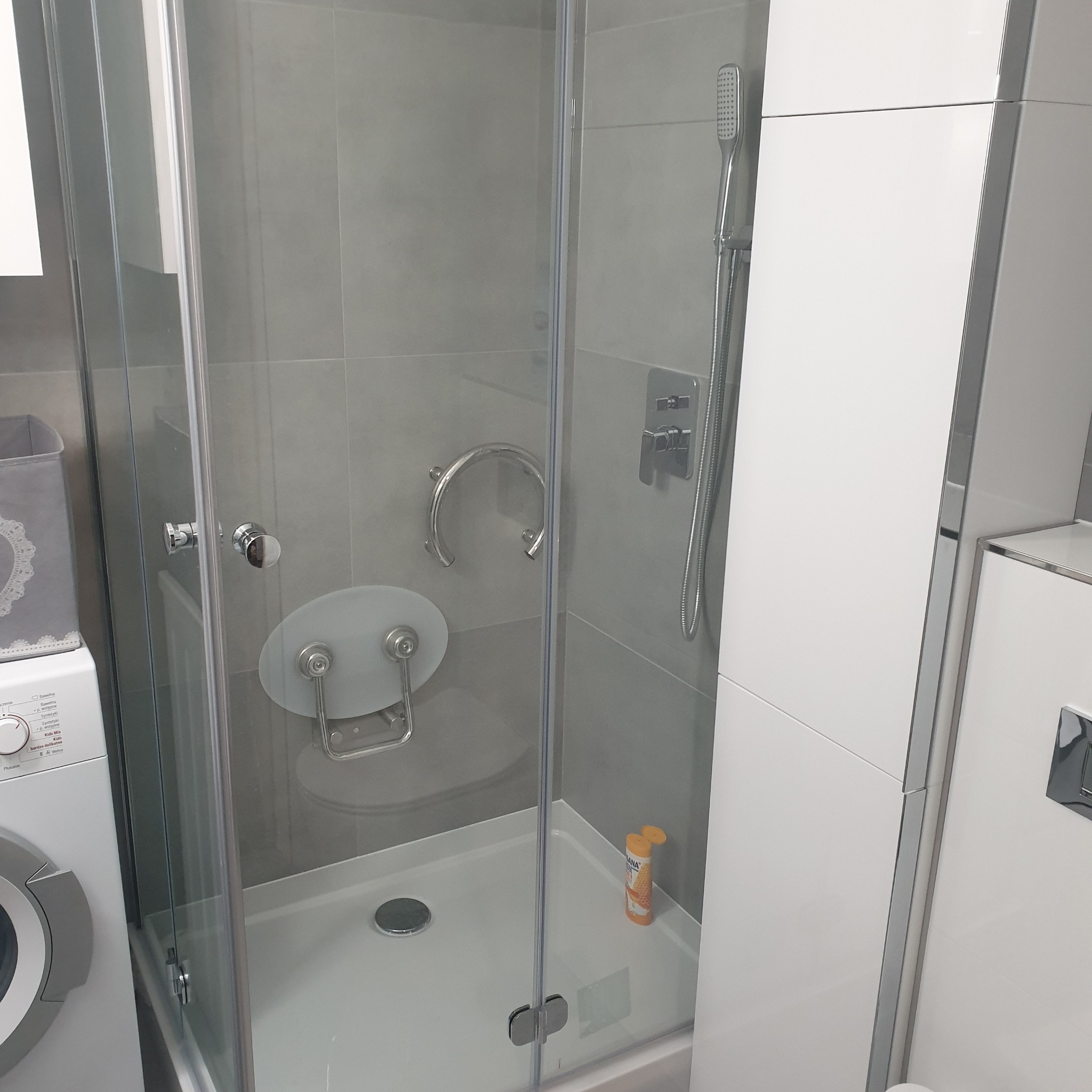 Szklana kabina prysznicowa z siedziskiem dla niepełnosprawnych i uchwytem, umieszczona w łazience obok pralki i białej szafki, z widocznymi szarymi płytkami na ścianie.