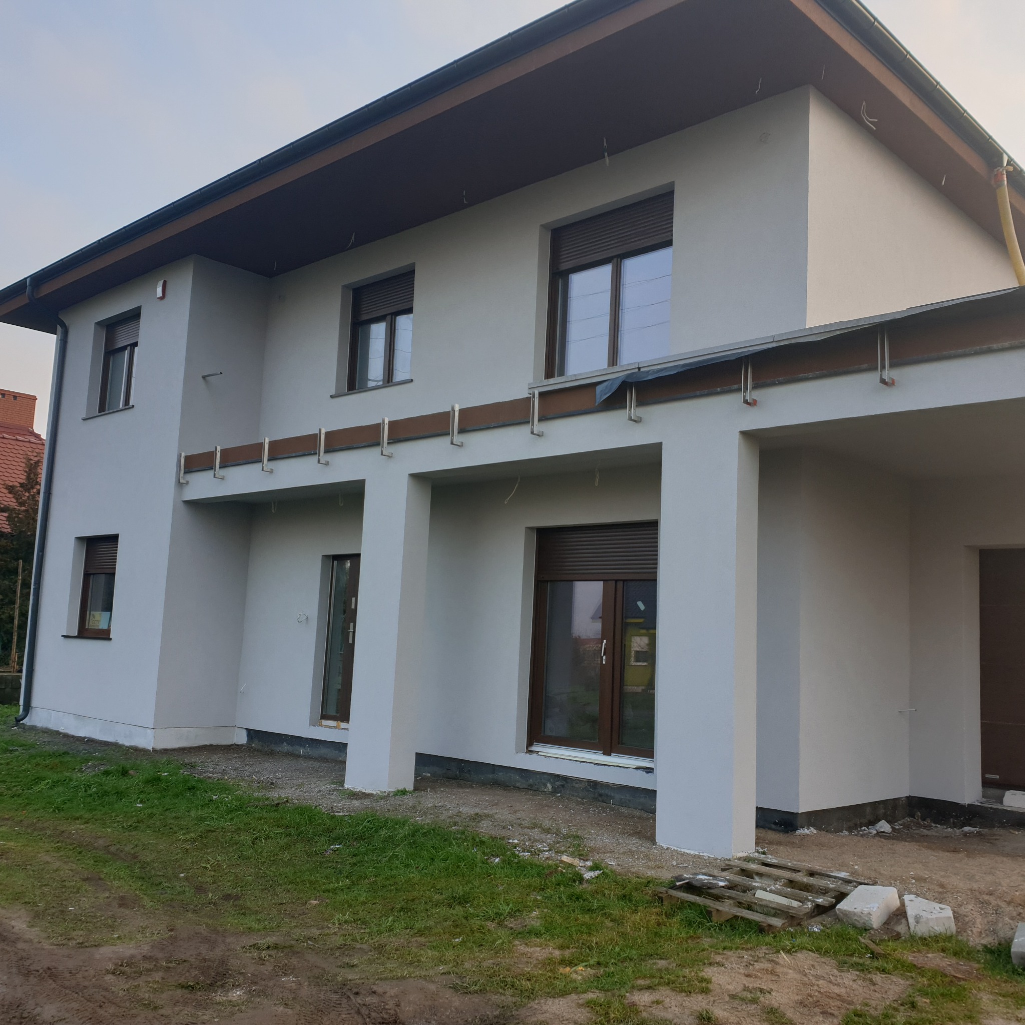 Nowoczesny dom jednorodzinny z jasną elewacją, brązowymi oknami i roletami zewnętrznymi, widoczne elementy konstrukcyjne balkonu i fragment terenu wokół domu w trakcie prac wykończeniowych.