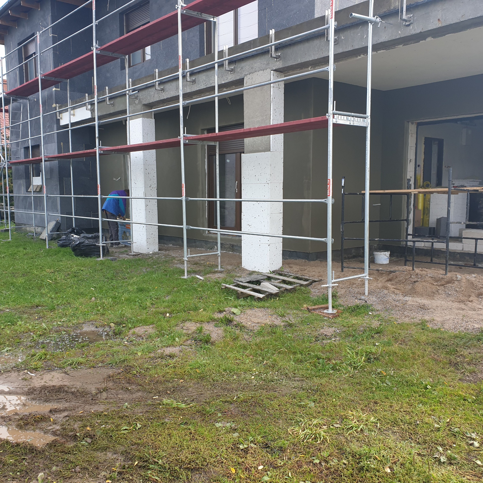 Prace ociepleniowe na elewacji domu jednorodzinnego z użyciem styropianu, widoczne rusztowania i pracownik w niebieskiej kurtce, teren wokół budynku częściowo błotnisty.