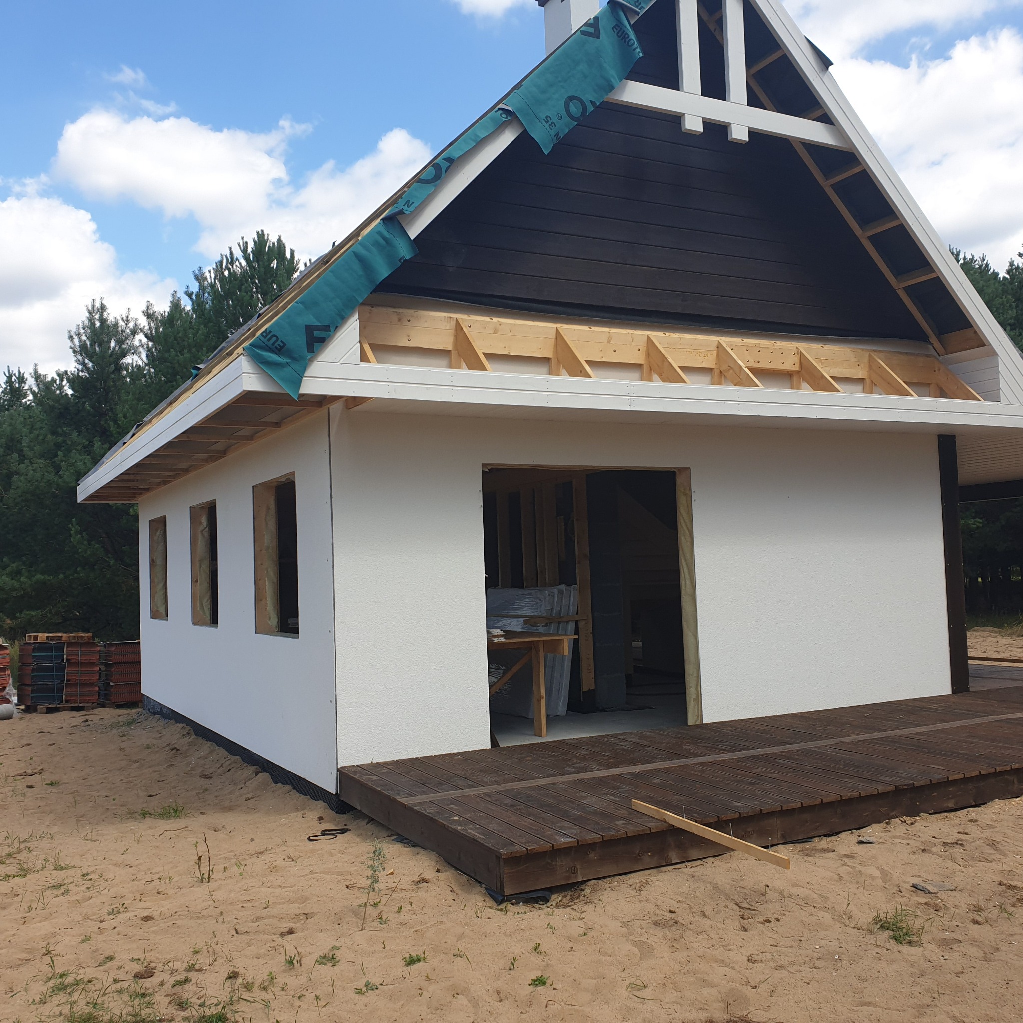 Dom w trakcie budowy: biała elewacja z fakturą, widoczne drewniane elementy konstrukcyjne dachu, brakujące okna, drewniany taras na piaszczystym podłożu, błękitne niebo z chmurami.