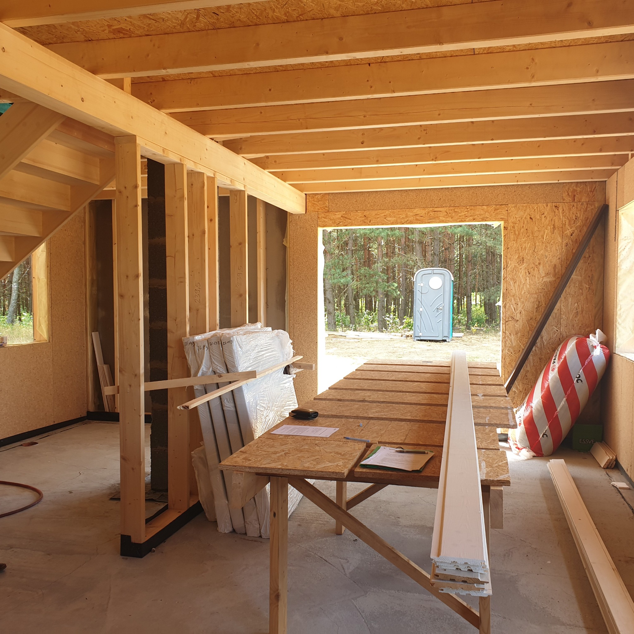 Wnętrze surowego domu w trakcie budowy, widoczne drewniane konstrukcje ścian i stropu, osb płyty, materiały izolacyjne w workach, tymczasowy stół z dokumentami, oraz toaleta przenośna widziana...