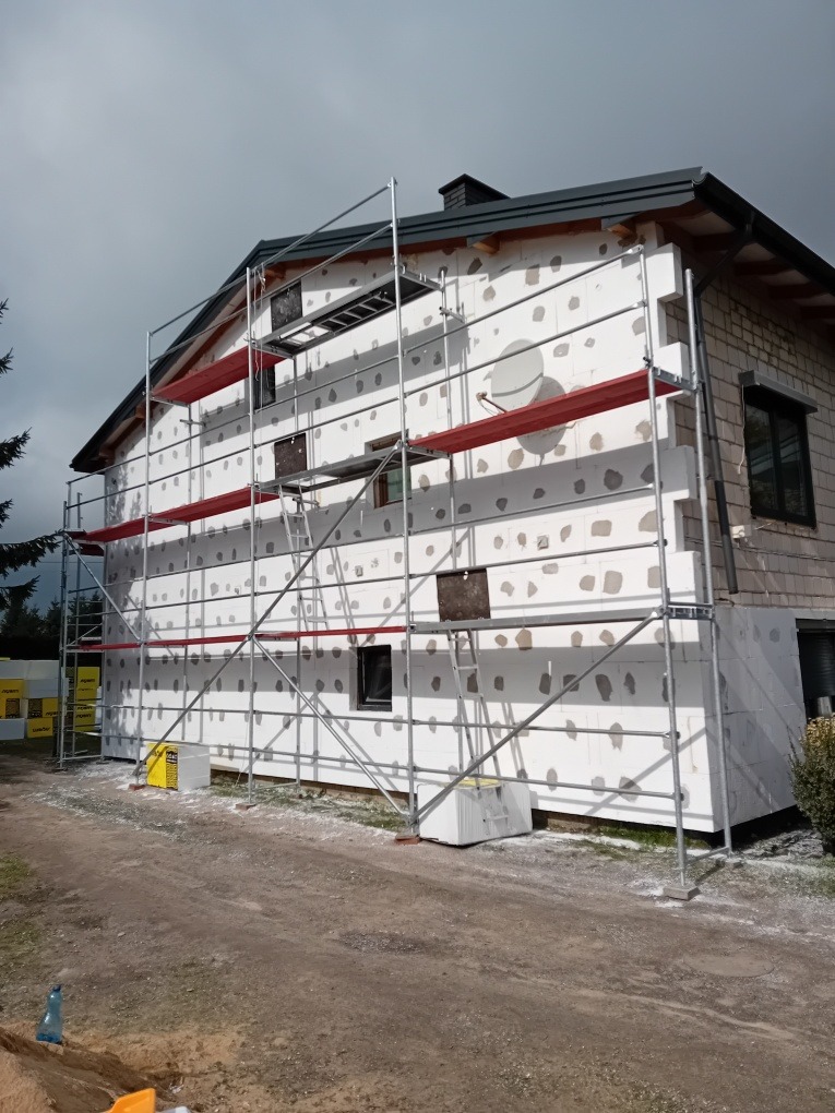 Dom jednorodzinny w trakcie ocieplania styropianem, widoczne rusztowanie i częściowo zaizolowana ściana szczytowa.