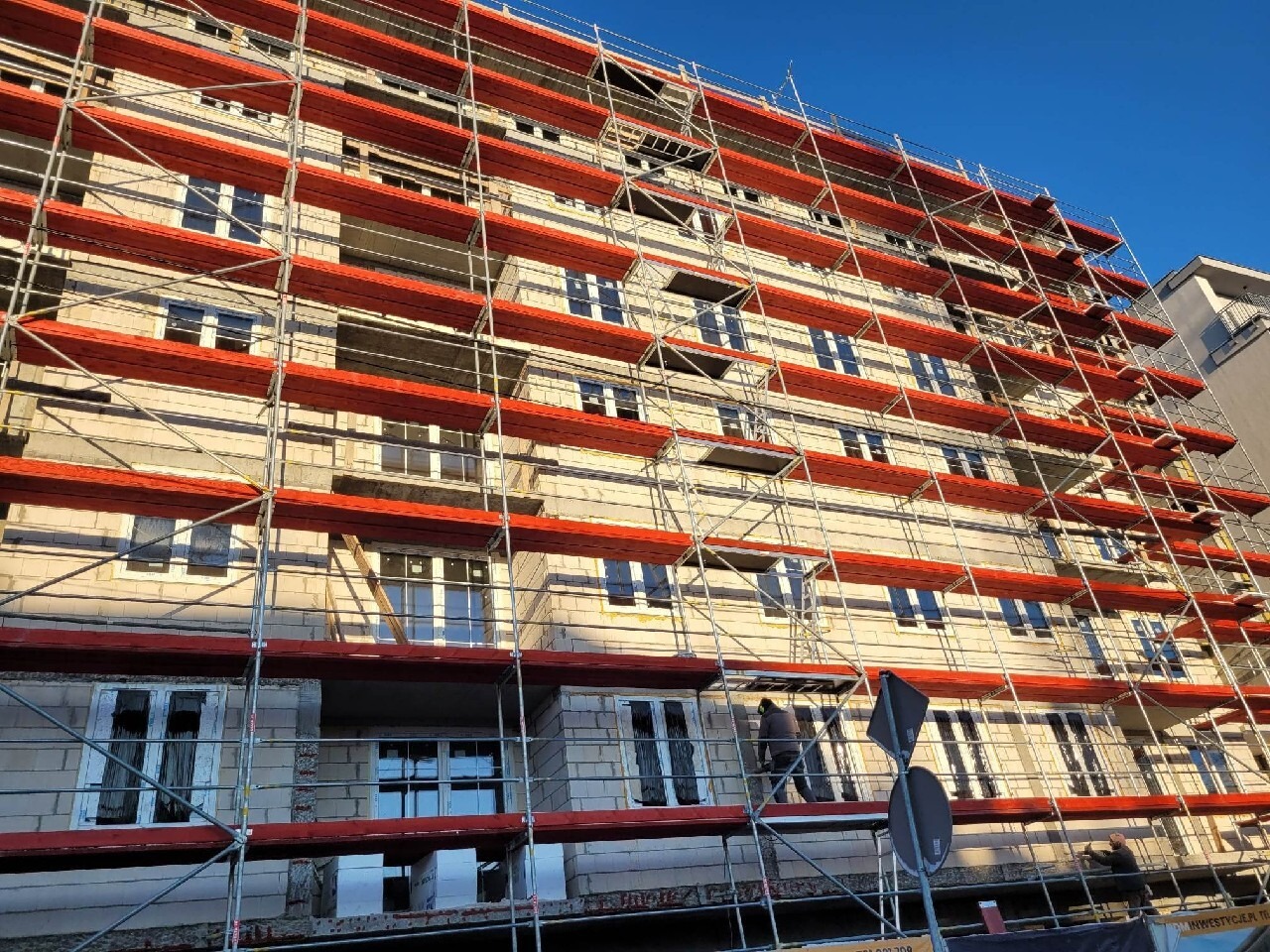 Wielopiętrowy budynek w trakcie montażu okien, otoczony rusztowaniem z czerwonymi zabezpieczeniami, widok z dołu na błękitne niebo.