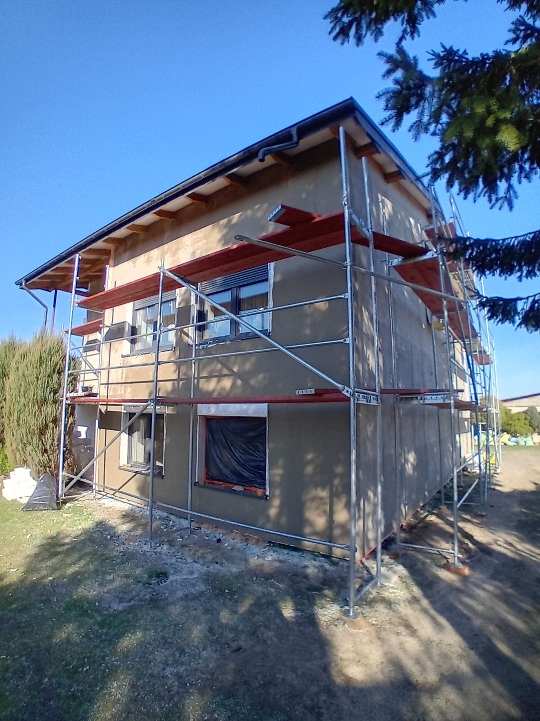 Dom jednorodzinny w trakcie prac elewacyjnych, otoczony rusztowaniem, z widocznym fragmentem okna zabezpieczonego folią i świeżo nałożonym tynkiem.