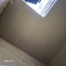 Świeżo nałożona, szara warstwa tynku na suficie, widoczna z perspektywy podłogi, z fragmentem okna w górnym rogu.