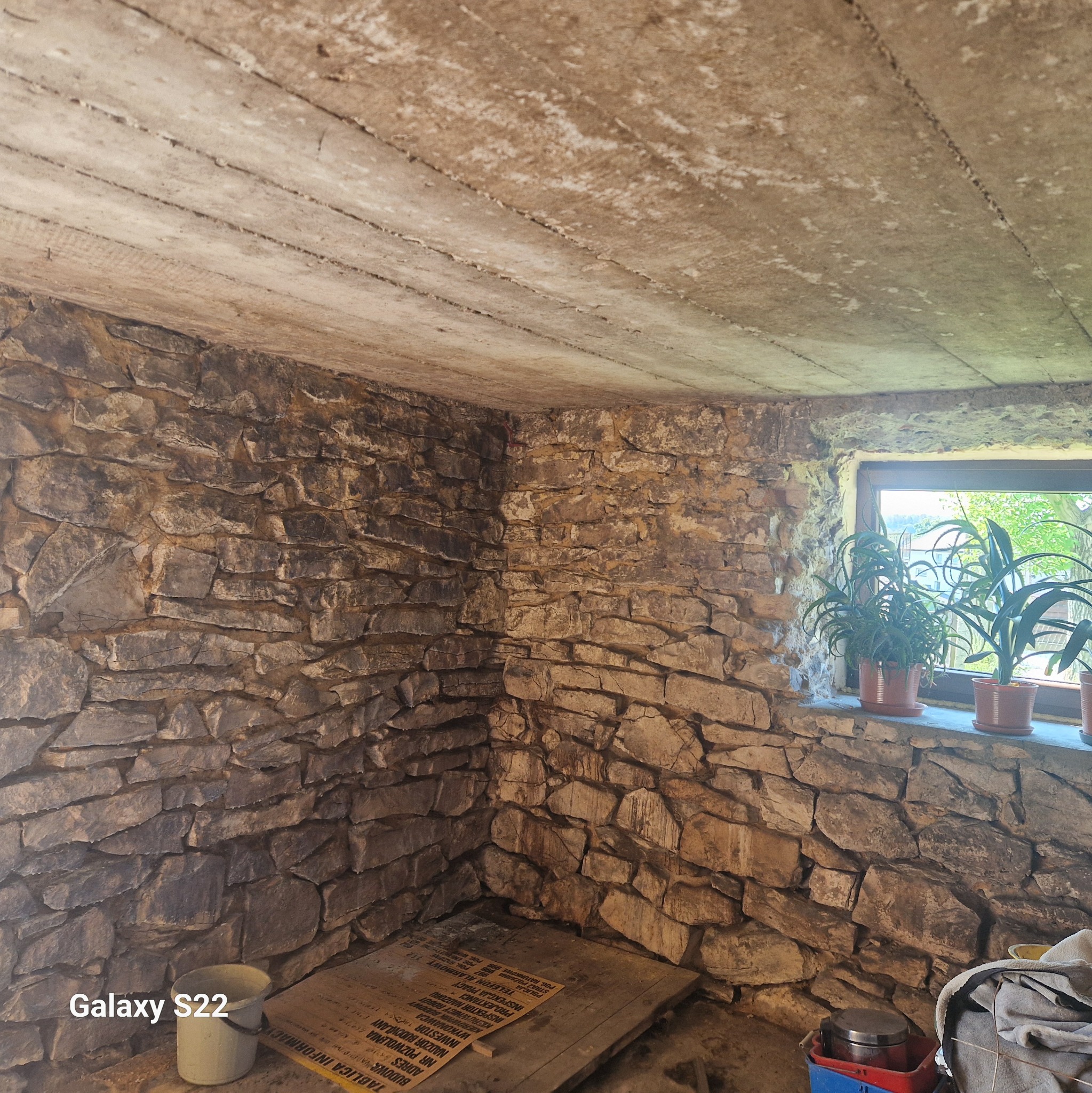 Surowe wnętrze piwnicy z kamiennymi ścianami, betonowym sufitem z widocznymi deskami szalunkowymi i oknem z roślinami doniczkowymi. W rogu widoczne wiadro i tablica informacyjna budowy.