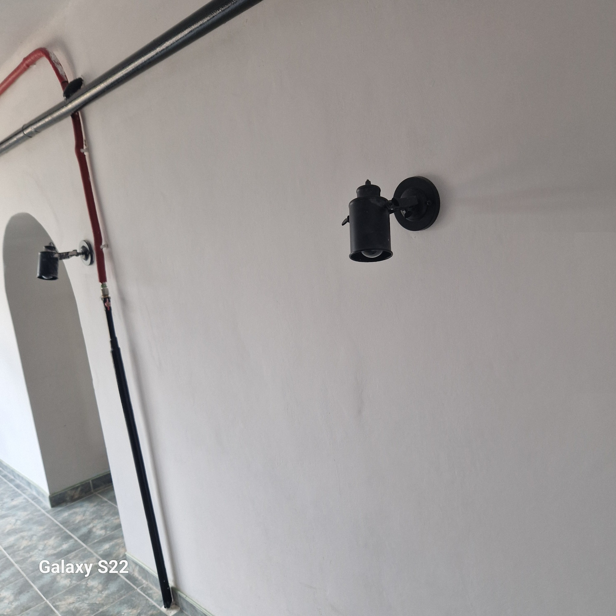 Czarna lampa ścienna w stylu industrialnym zamontowana na gładkiej, białej ścianie, widoczne rury instalacyjne biegnące wzdłuż sufitu i korytarz z szarą podłogą w tle.