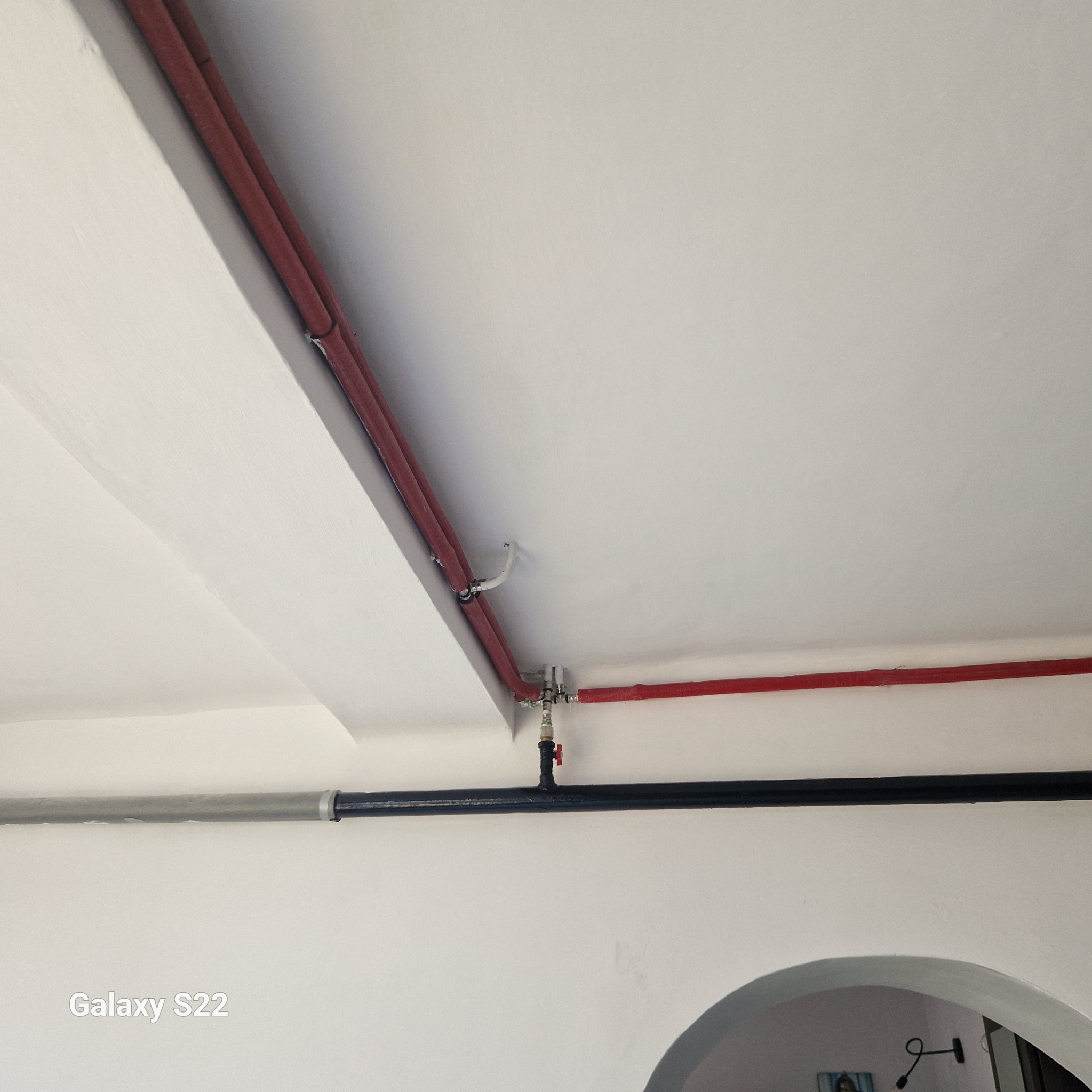 Biały sufit z instalacją wodną: czerwone rury prowadzone wzdłuż krawędzi sufitu i czarna rura z zaworem, widoczny fragment łuku w ścianie.