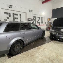 ŻELAZO TEAM SPÓŁKA Z OGRANICZONĄ ODPOWIEDZIALNOŚCIĄ - Dwa samochody Audi A4 w warsztacie, jeden srebrny kombi z tyłu, drugi czarny sedan z otwartą maską, widoczny płyn do spryskiwaczy, na ścianie półki z produktami Fischer i dekoracje z szyb.