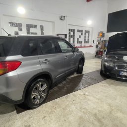 ŻELAZO TEAM SPÓŁKA Z OGRANICZONĄ ODPOWIEDZIALNOŚCIĄ - Szary samochód Suzuki Vitara po umyciu, z widocznymi kroplami wody, zaparkowany obok czarnego Audi A4 z otwartą maską w jasnym garażu z półkami pełnymi środków czyszczących.