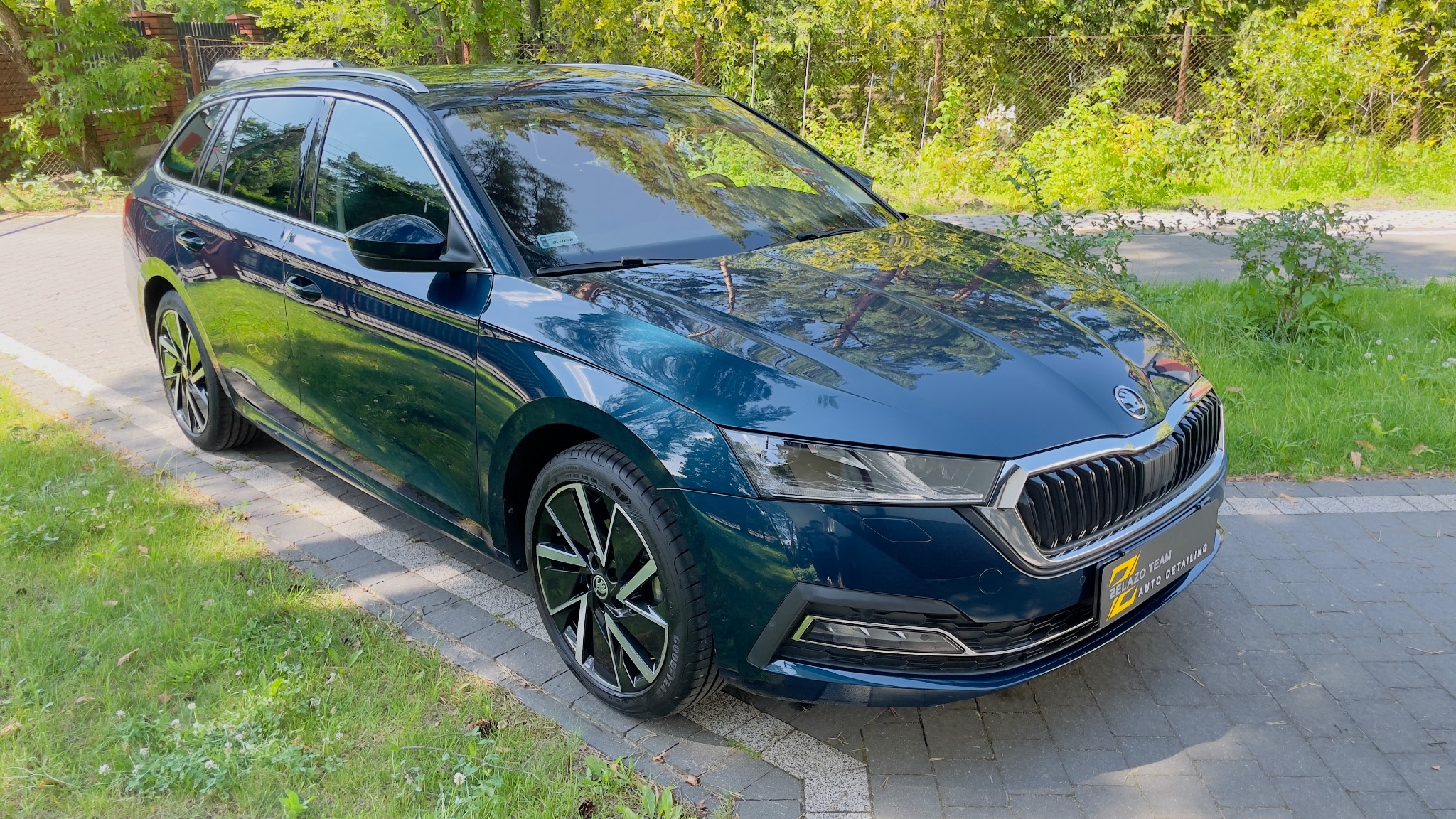 Błyszczący, granatowy samochód kombi Skoda Octavia po detailingu, zaparkowany na kostce brukowej, z widocznym logo firmy detailingowej na przednim zderzaku.