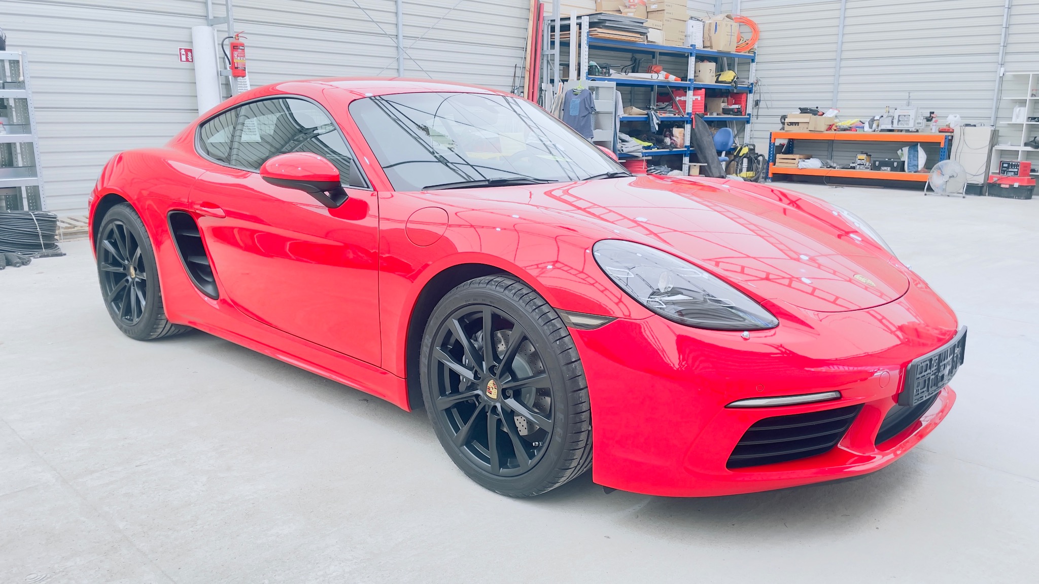 Czerwone Porsche 718 Cayman z czarnymi felgami, zaparkowane w jasnym garażu z regałami w tle. Widoczne detale karoserii i oświetlenie.