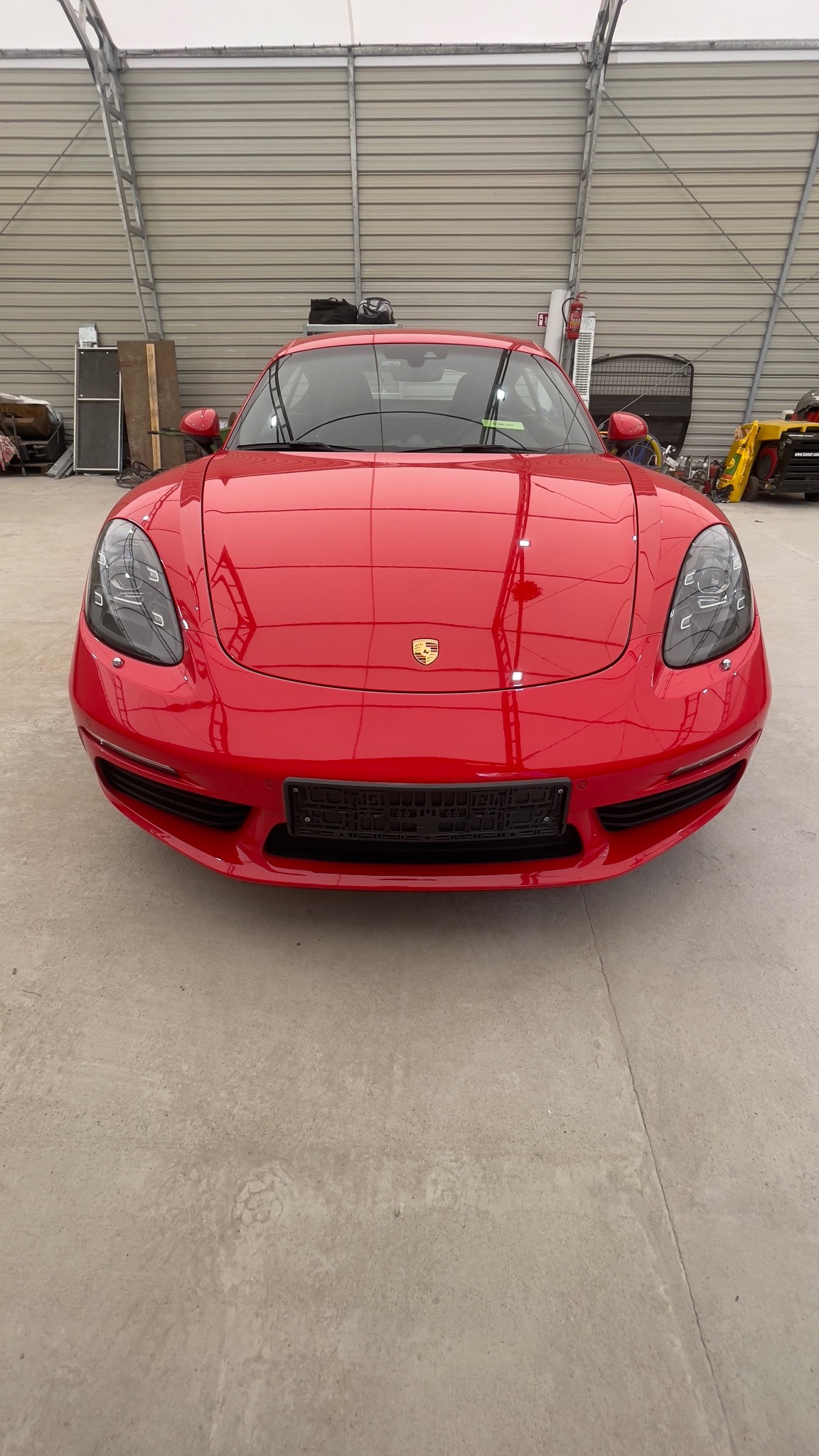 Czerwony sportowy samochód Porsche, widok z przodu, zaparkowany w przestronnym garażu z betonową podłogą i metalową konstrukcją dachu.