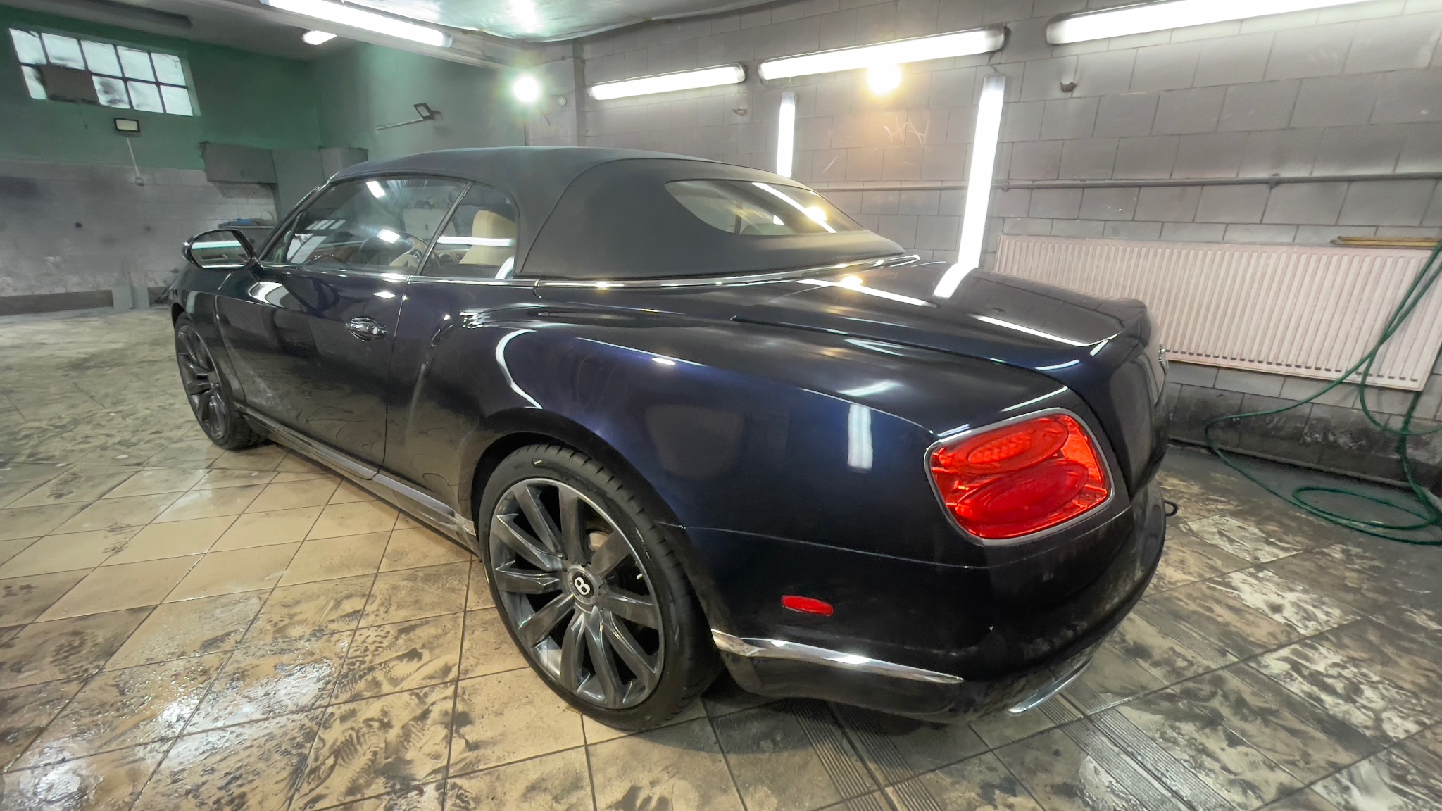 Granatowy kabriolet Bentley Continental z czarnym dachem, po myciu, widoczny z tyłu pod kątem, na mokrej posadzce warsztatu z oświetleniem jarzeniowym.
