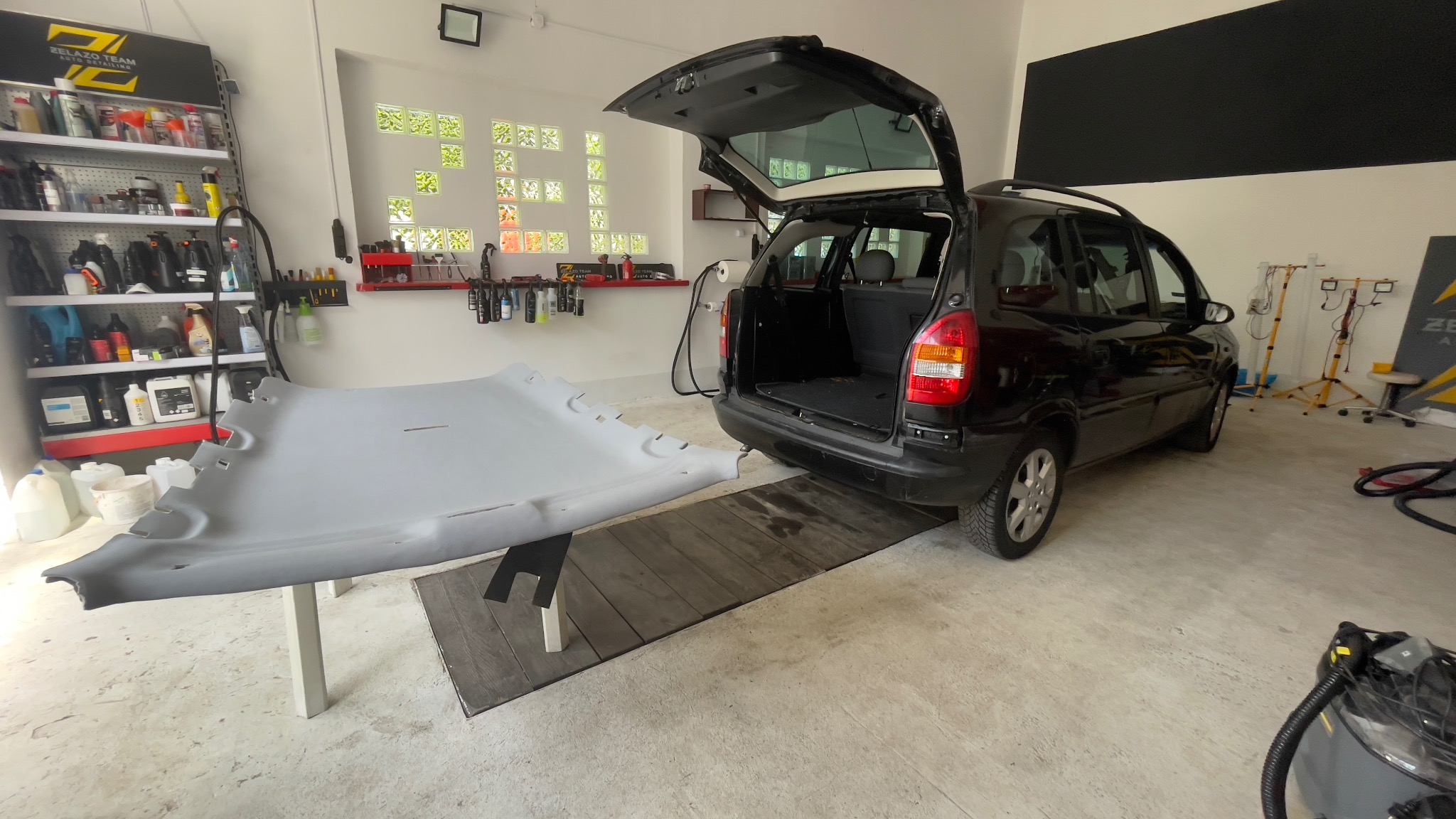 Demontaż podsufitki w czarnym kombi Opel Astra, widoczne narzędzia i środki czyszczące na półkach w warsztacie.