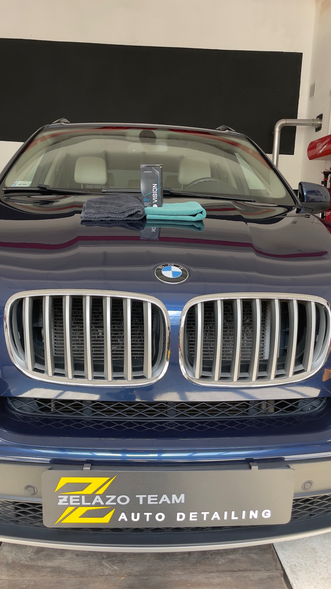 Niebieski SUV BMW z produktem do pielęgnacji lakieru Vision, szarą ściereczką z mikrofibry i turkusową szmatką na masce, widoczne logo Żelazo Team Auto Detailing na tablicy rejestracyjnej.