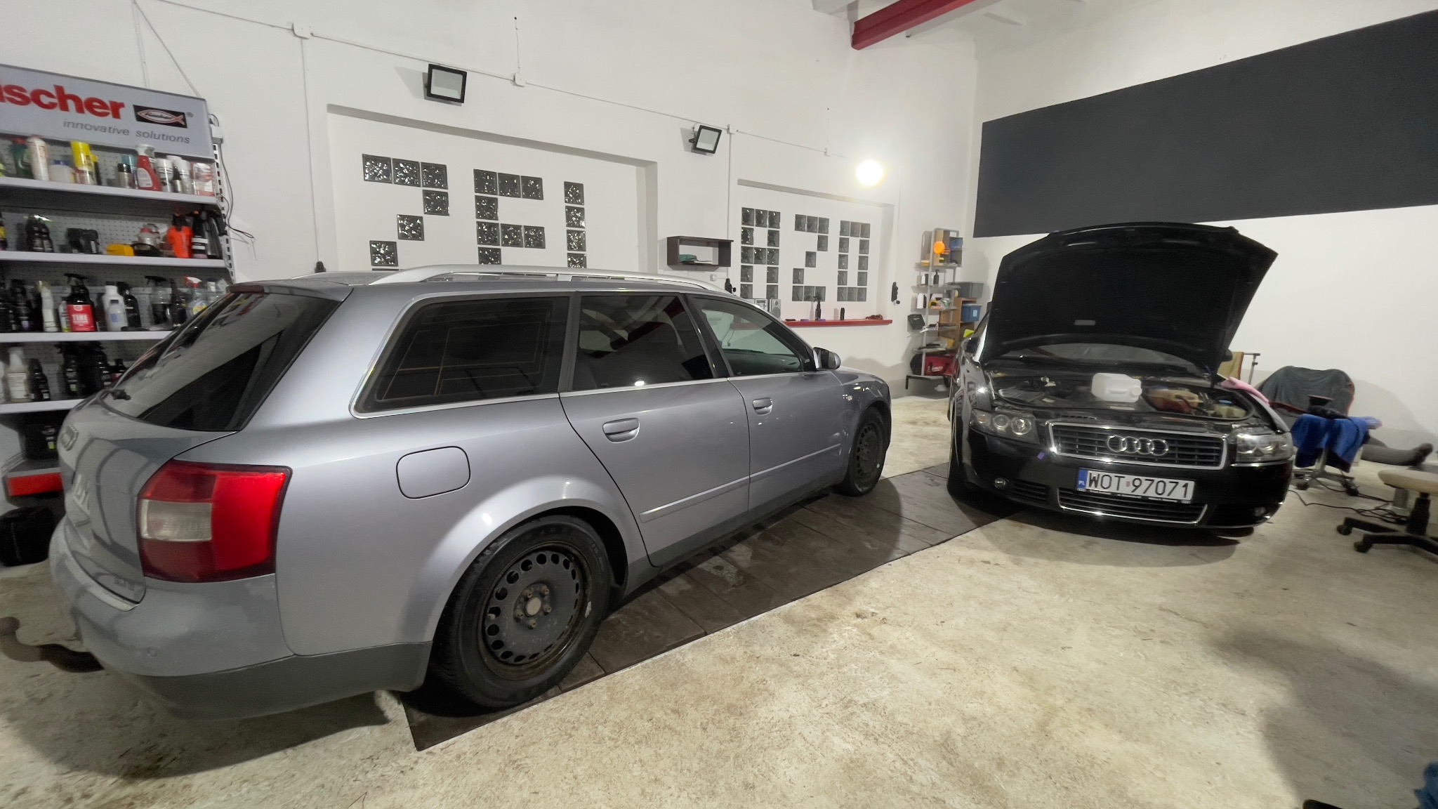 Dwa samochody Audi A4 w warsztacie, jeden srebrny kombi z tyłu, drugi czarny sedan z otwartą maską, widoczny płyn do spryskiwaczy, na ścianie półki z produktami Fischer i dekoracje z szyb.