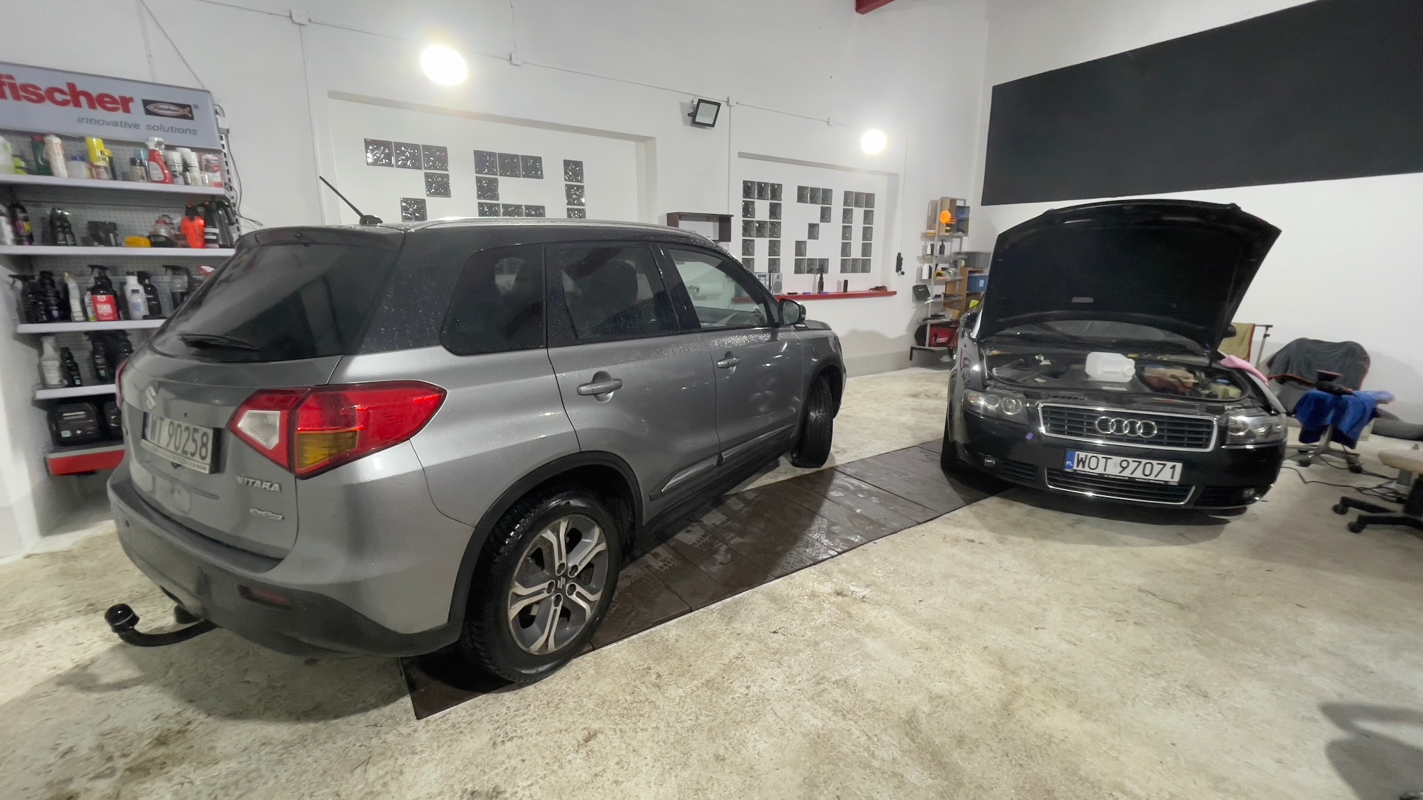 Szary samochód Suzuki Vitara po umyciu, z widocznymi kroplami wody, zaparkowany obok czarnego Audi A4 z otwartą maską w jasnym garażu z półkami pełnymi środków czyszczących.