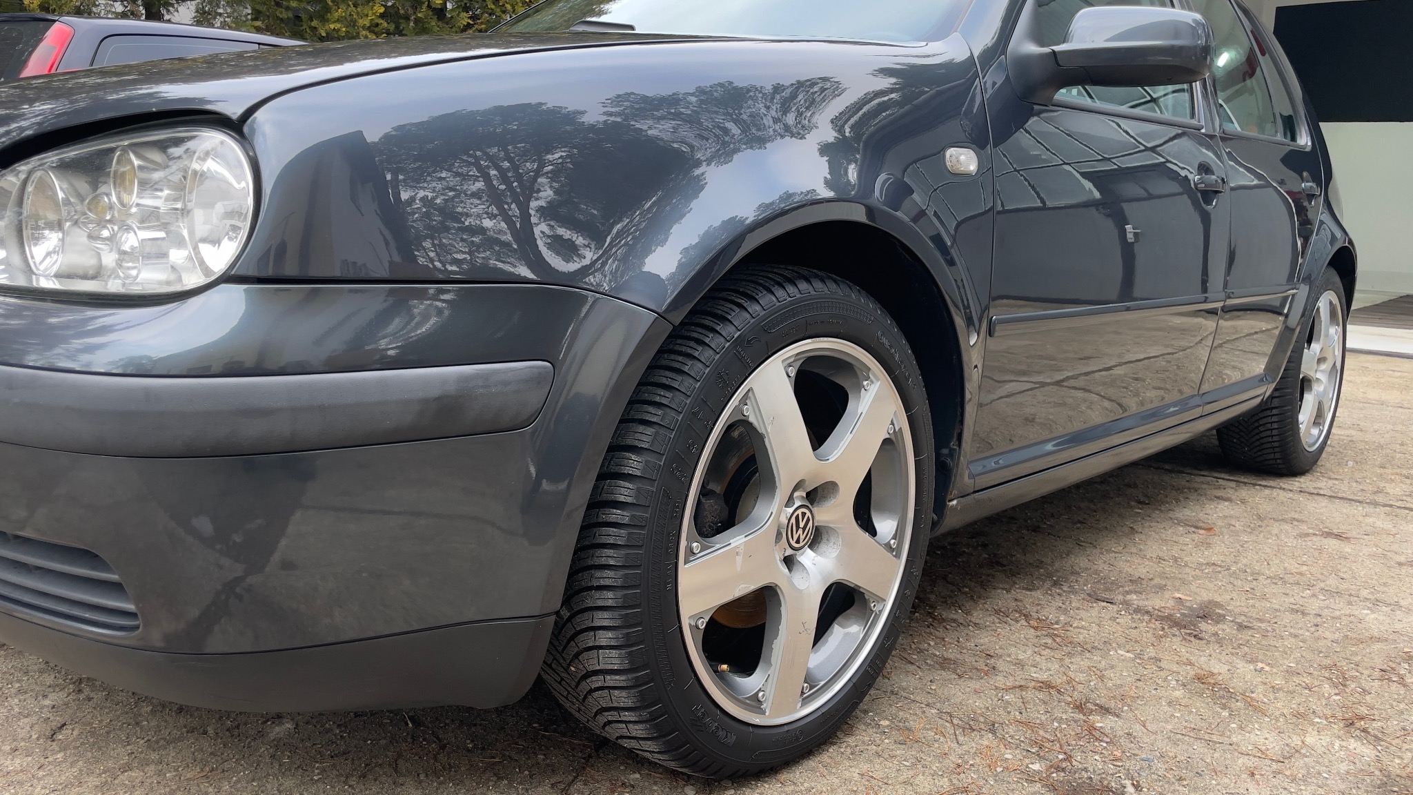 Szary Volkswagen Golf z srebrnymi felgami, widoczne opony zimowe z bieżnikiem, karoseria odbija drzewa.