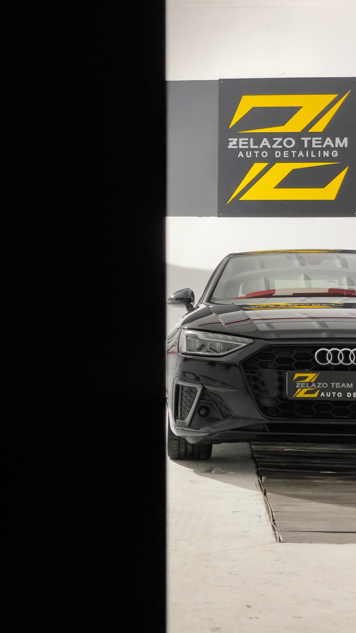 Częściowy widok czarnego Audi A4 w studiu auto detailingu z logo firmy Zelazo Team Auto Detailing na ścianie.