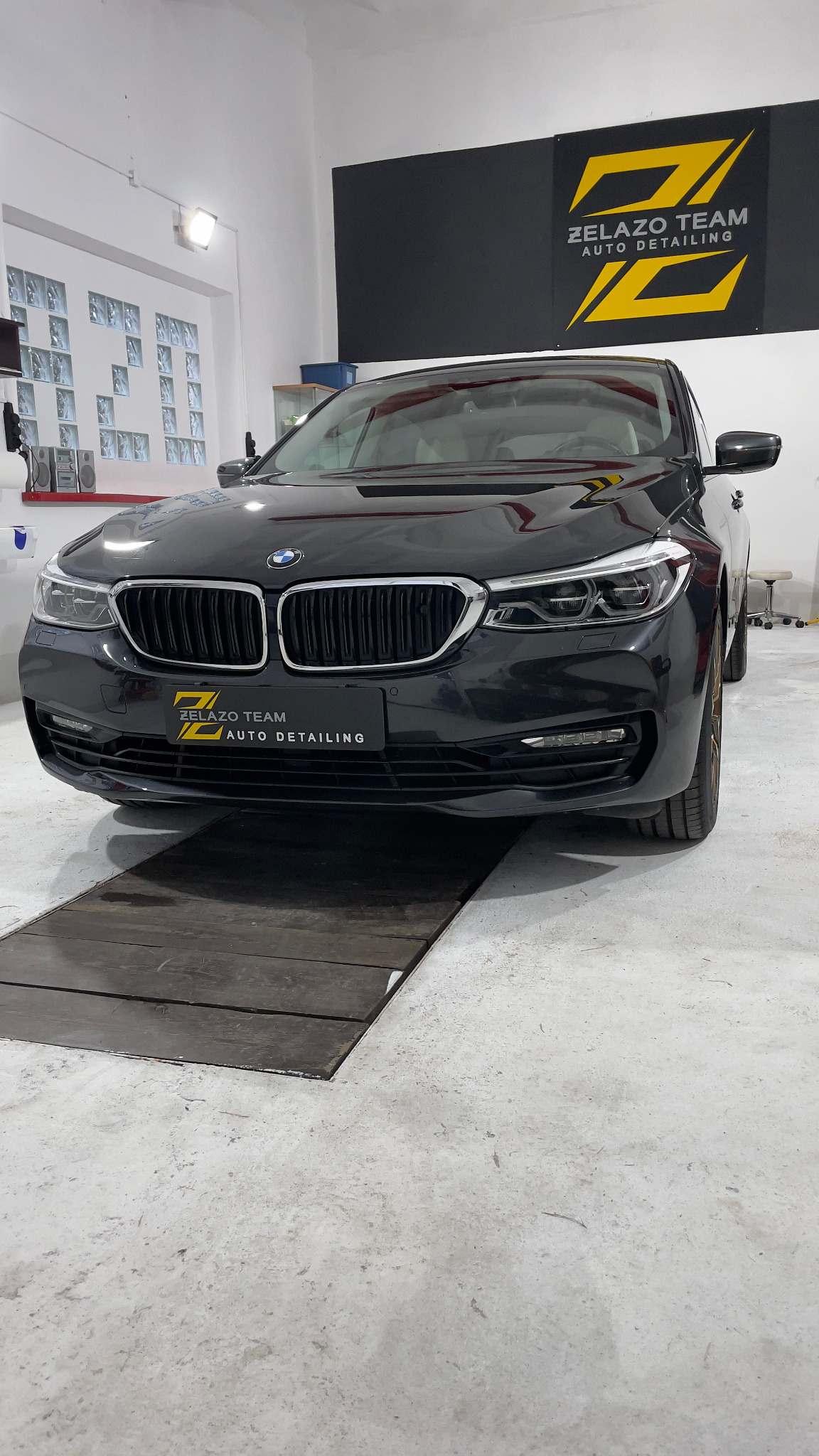 Czarny samochód BMW po detailingu, zaparkowany na drewnianej platformie w jasnym warsztacie z logo firmy 'Zelazo Team Auto Detailing' widocznym na ścianie i tabliczce na pojeździe.
