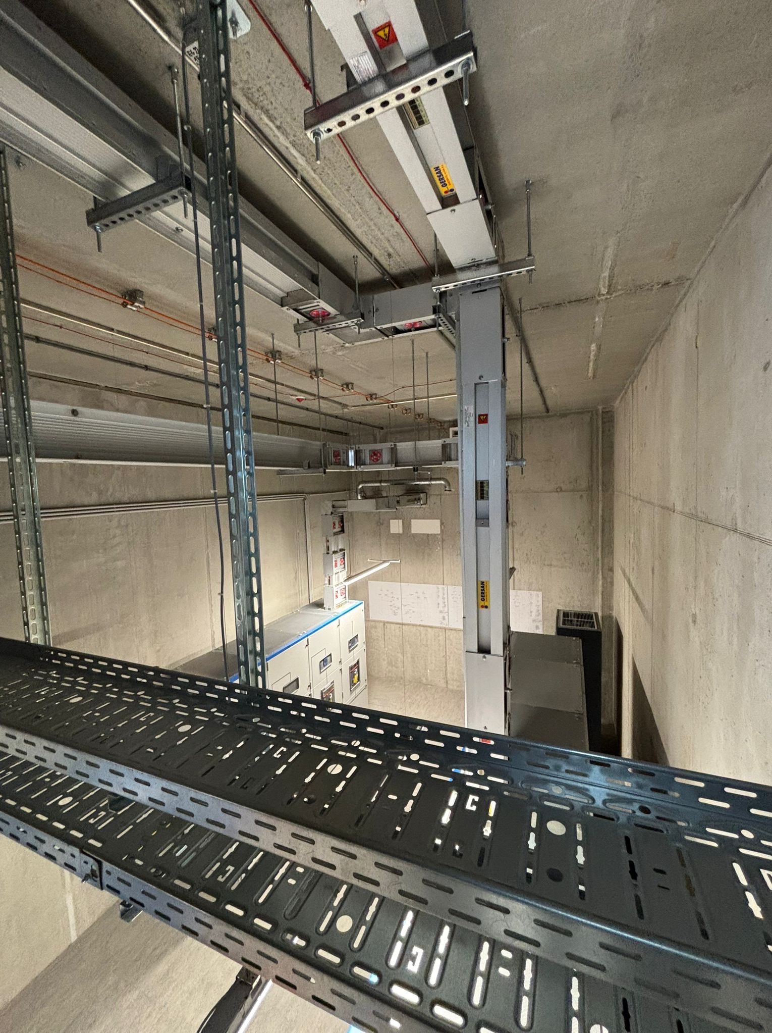 Widok z góry na koryto kablowe, szafę sterowniczą i instalacje pod sufitem w surowym, betonowym pomieszczeniu technicznym.