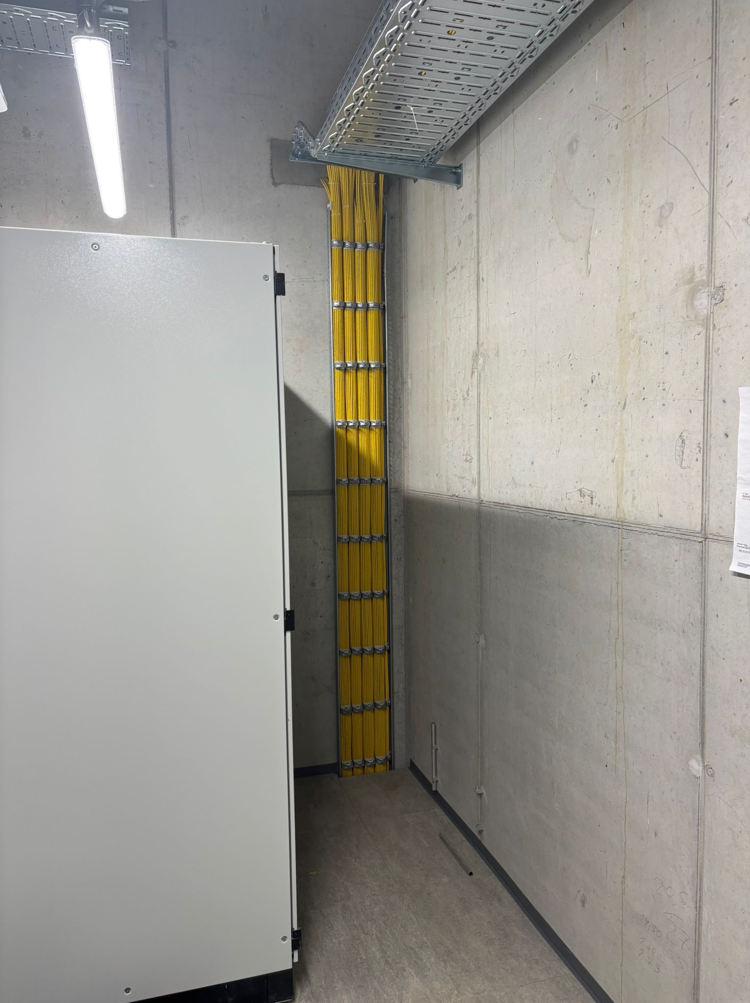 Żółte kable ułożone w pionowym korycie, prowadzone od sufitu do podłogi w surowym, betonowym pomieszczeniu technicznym, obok szafy sterowniczej.