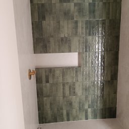 Marcin Machowski MarDom - Ściana wykończona błyszczącymi, podłużnymi, zielonymi płytkami w łazience. Widoczna wnęka i złota bateria. Minimalistyczny odpływ liniowy w podłodze.